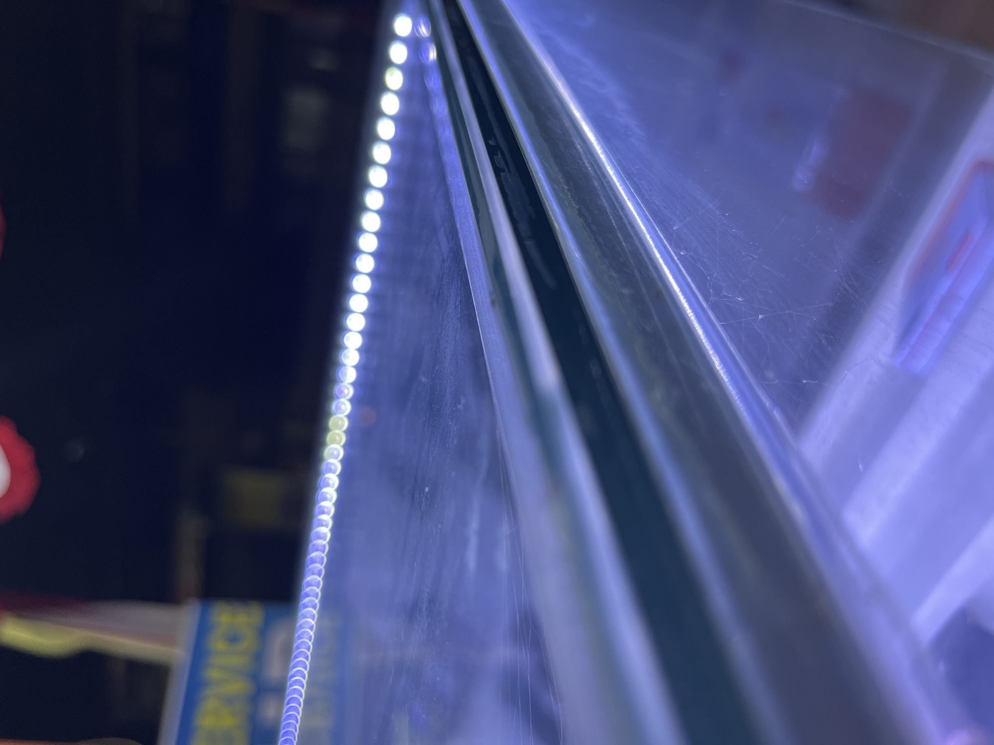 The image captures a close-up, low-angle view of a glass surface illuminated by a string of white LED lights. The lights are positioned along the edge of the glass, creating a reflective glow. The background is dark and out of focus, with a hint of a blue sign that appears to read "SERVICE." There are some faint lines or smudges visible on the glass surface. The overall scene suggests an indoor setting, potentially a display case or a shelf, in an urban environment, possibly Central Jakarta, Indonesia, given the presence of the "SERVICE" sign, which is common in commercial establishments. There are no people or discernible interactions visible in the image.