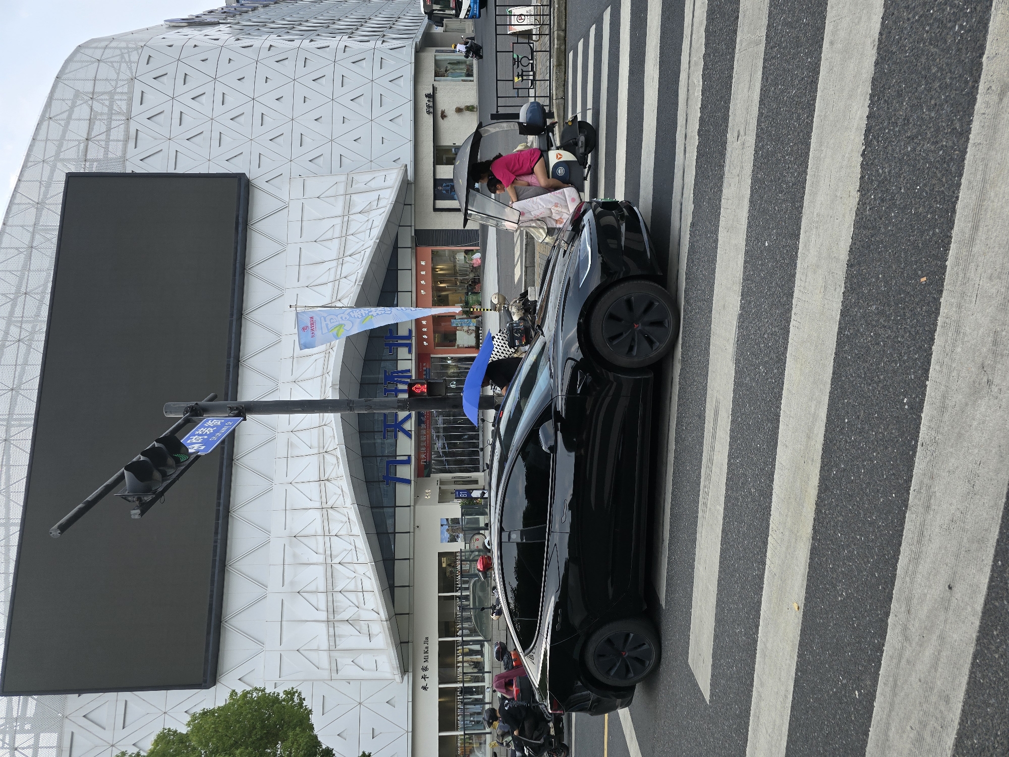 In Shangcheng District, China, a black Tesla is parked on a pedestrian crossing in front of a modern building with a distinctive geometric facade. The building's exterior features a large, dark digital screen and a white, textured surface composed of triangular patterns. A traffic light with a green signal is visible, indicating that vehicles may proceed.
Several people are present around a small, covered tricycle, with one person in a pink top and shorts leaning into the vehicle, possibly attending to a child lying on a blanket inside. The scene appears to be in daylight, and the weather is likely clear or partly cloudy.
Various signs with Chinese characters are visible on the building, including "米卡家 Mi Ka Jia" and "九天环北服装城 波函". A blue umbrella is also partially visible, suggesting it might be a sunny day or there's a possibility of rain. Several motorbikes are parked or moving in the background, typical of urban street scenes in China. The overall impression is a bustling urban environment with everyday activities taking place.