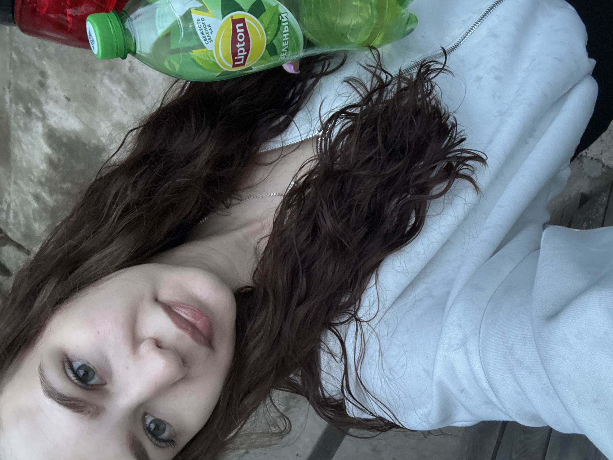 A woman is visible in a selfie-style image. She has brown hair, grey eyes, and light skin. She wears a white jacket with a zipper. Two plastic bottles, one with a green lid and the other with a red lid, are positioned near her neck. The green-lidded bottle has a "Lipton" label. The background appears to be an outdoor setting. Information on Nizhnyaya Gar', Russia is not discernible from the provided image.