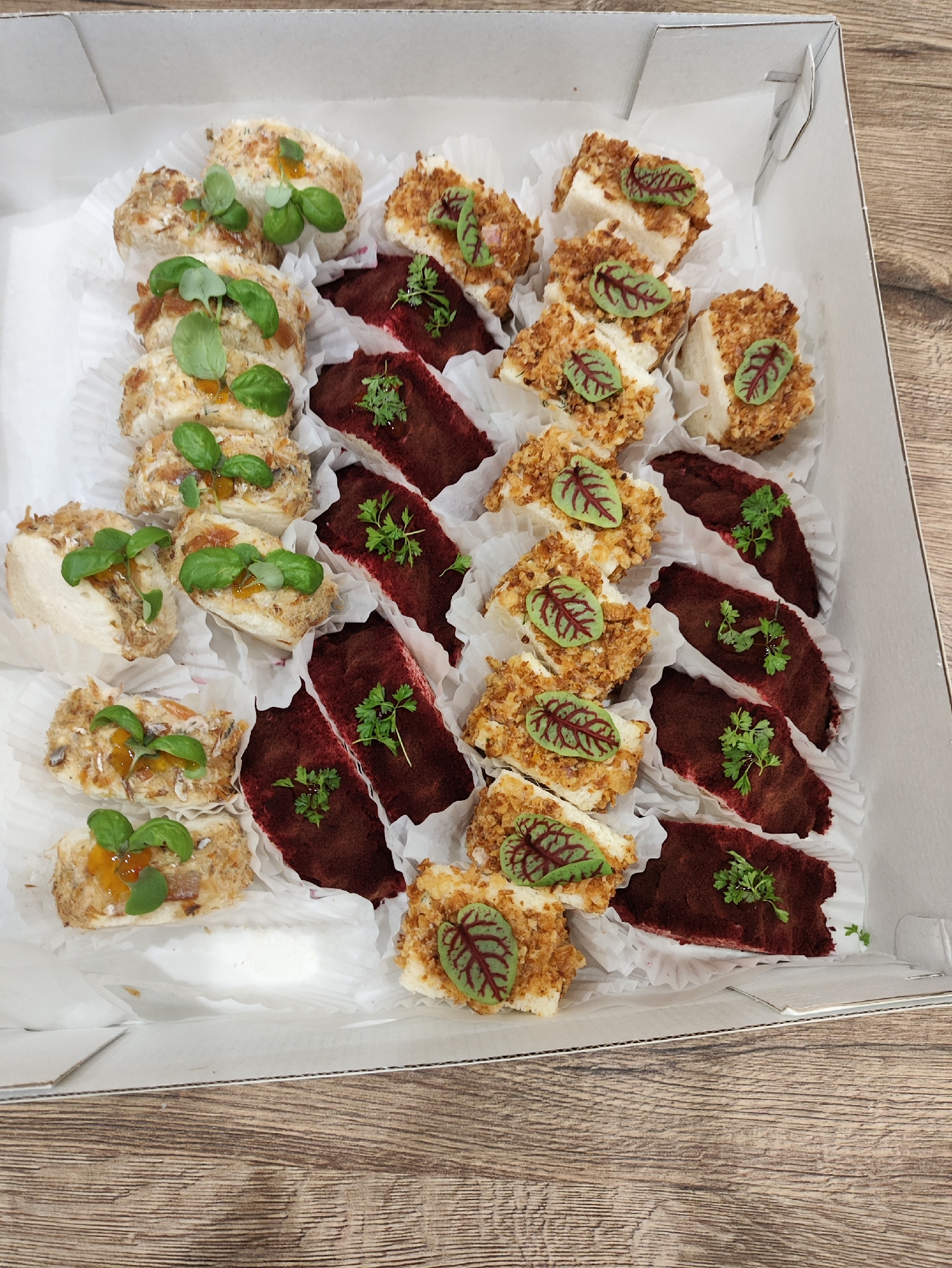 A white cardboard box contains an assortment of small, bite-sized food items arranged on white paper liners. The food appears to be appetizers or canapés.
There are two main types of items visible. One type is light-colored, possibly cream cheese or a similar base, covered in a crumbly topping and garnished with green herbs and some small pieces of orange. Another type is a dark red, rectangular item, possibly a pâté or terrine, also garnished with green herbs. Some of these red items are topped with small, decorative red and green leaves.
The food is arranged in rows within the box. The box itself is positioned on a wooden surface, likely a table, with visible wood grain. The lighting suggests it is daytime, and the setting appears to be indoors, possibly a catering or food preparation environment. There is no text visible in the image. The overall impression is of a prepared selection of appetizers.