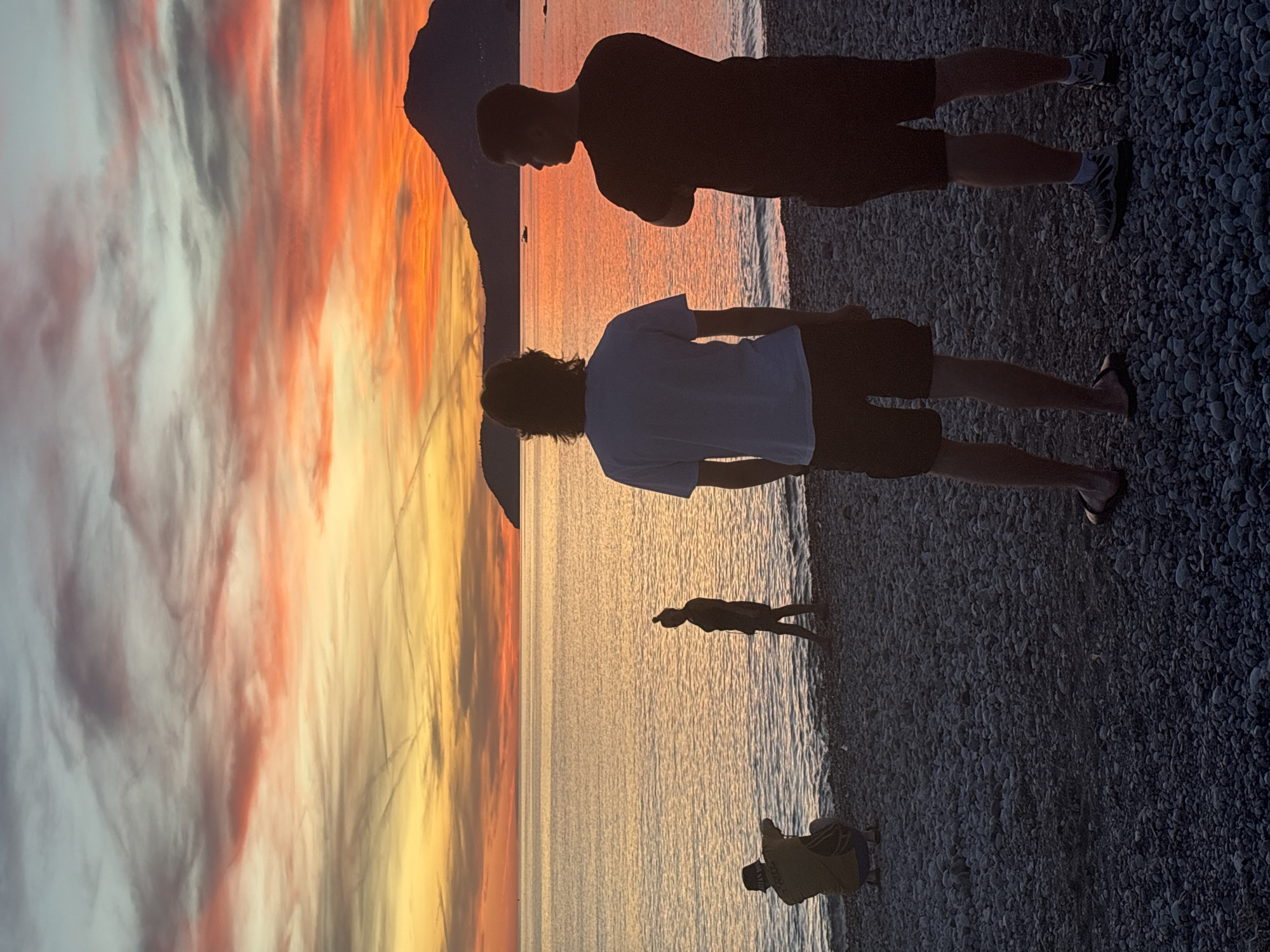 The image depicts a sunset scene on a pebble beach, likely in the city of Bar, Montenegro, given the presence of a distinctive mountain profile in the distance characteristic of the region. The sky is filled with vibrant colors ranging from deep orange and pink to yellow and light blue, indicating either sunrise or sunset. Wispy clouds are scattered across the sky, some streaked with contrails.
In the foreground, four individuals are visible. On the left, a person in a yellow top and dark bottoms is crouched down, facing the water, possibly taking photographs or observing something. Slightly further back and to the right, a silhouetted figure stands near the water's edge, facing away from the camera. Dominating the center right of the frame are two young men, also in silhouette, standing with their backs to the camera. The one on the left wears a white t-shirt and dark shorts, while the one on the right wears a dark t-shirt and dark shorts. They are looking out towards the sea and the horizon.
The sea itself is calm, reflecting the colors of the sky. The beach is composed of small, grey pebbles. In the background, a mountain range, a prominent peak with a structure on top, rises from the sea, suggesting a coastal landscape typical of Bar. A small boat is visible on the water in the mid-ground. The overall atmosphere is serene and picturesque, capturing a moment of tranquil observation of nature's beauty.