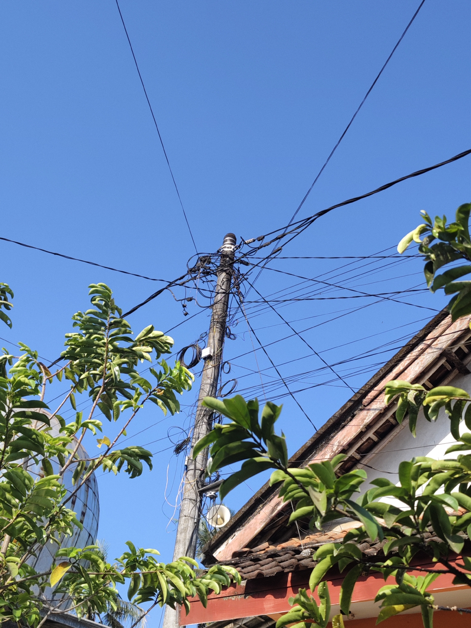 In Bangsalsari, Indonesia, this image captures a close-up view of a utility pole laden with a chaotic network of electrical and telecommunication wires against a clear blue sky. The pole, positioned centrally in the frame, is densely wrapped with numerous black wires, some thick and others thin, indicating a complex and possibly unorganized infrastructure.
To the right of the pole, the edge of a building's roof is visible, characterized by terracotta tiles and a white wall. Green foliage from nearby trees frames the scene on the left and right sides, their leaves vibrant and slightly blurred, suggesting they are closer to the camera. The overall environment appears to be an outdoor, urban or semi-urban setting with evidence of residential or commercial structures. The presence of the utility pole and the building suggests a populated area. No people are visible in the image. The lighting indicates it is daytime, likely with direct sunlight casting shadows that are not prominently featured.
