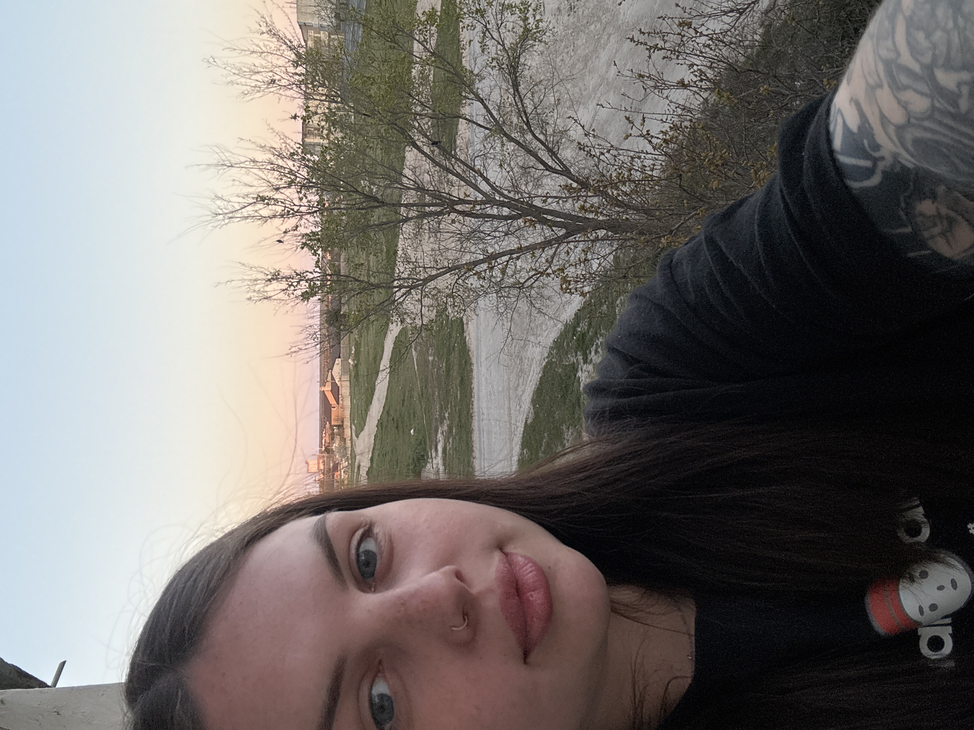 The image is a selfie of a young woman taken outdoors in Chornomorskoe, Ukraine, during what appears to be either sunset or sunrise. The woman, who is the primary subject, is positioned in the foreground, looking directly at the camera with a neutral to slightly serene expression. She has long dark hair, blue eyes, fair skin with some freckles, and wears a septum piercing. Her lips are full and slightly parted.
She is dressed in a dark, likely black, long-sleeved shirt. On her shirt, near her right shoulder (visible on the right side of the image), there is a white and red-capped character resembling an "Among Us" crewmate, with partial text "adi" visible beside it, suggesting an Adidas brand. Her right arm, which is partially visible, is covered in extensive, intricate tattoos. A light-colored concrete or stone surface is visible in the bottom-left corner, possibly a ledge she is leaning against.
The background reveals a transitional landscape with dirt paths winding through areas of grass and bare earth. Several trees, some with sparse green leaves budding and others still mostly bare, are scattered across the mid-ground. In the far distance, buildings indicating a town or settlement are visible. The sky transitions from a soft light blue on the left to delicate pink and orange tones on the right, characteristic of dawn or dusk. The overall setting suggests a somewhat rural or suburban periphery of Chornomorskoe.
