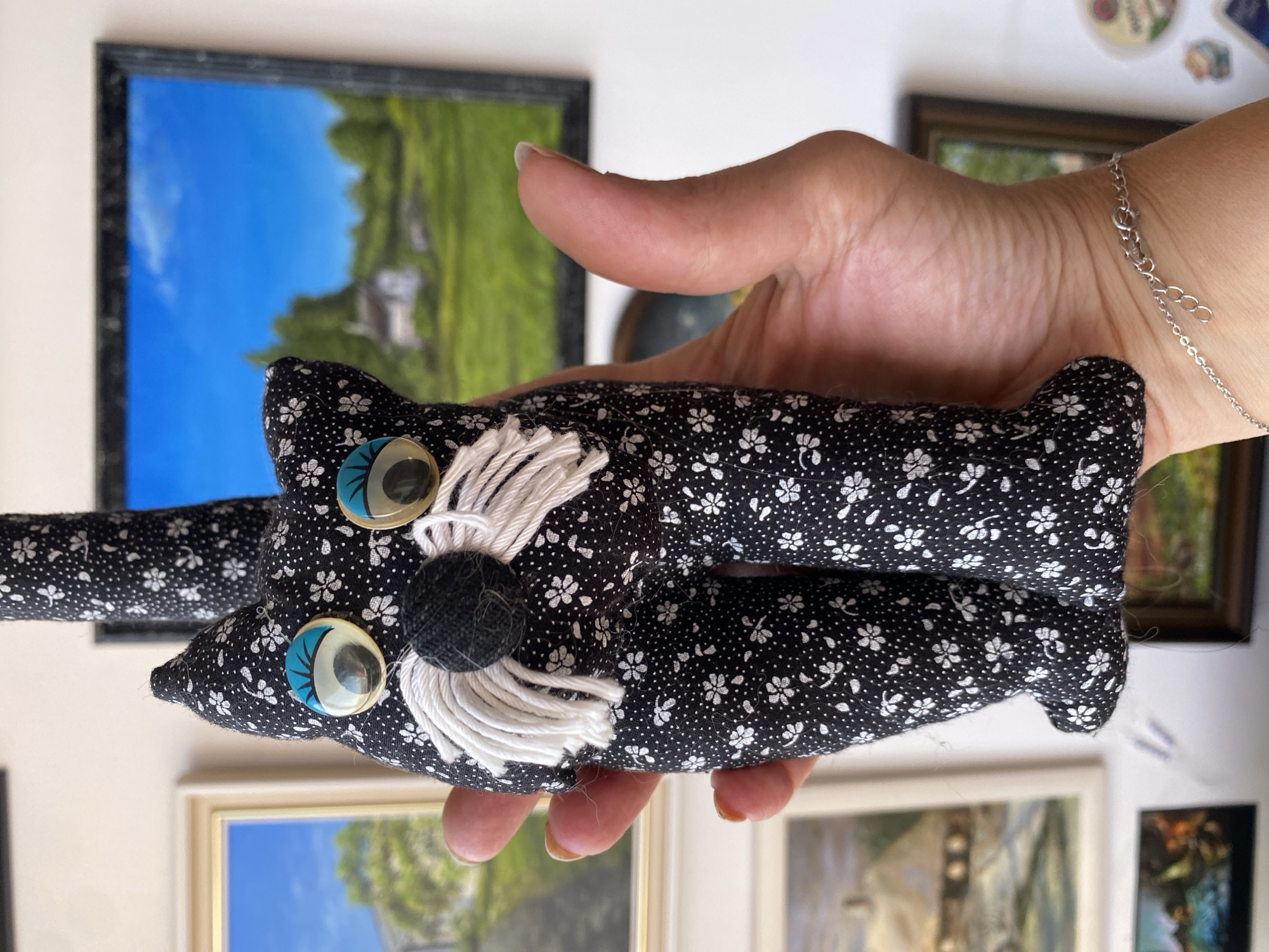 The scene is situated indoors in the city of Bílina, Czechia. A human hand, positioned in the foreground, holds a fabric plush toy designed to resemble a cat. The plush toy is approximately 20-25 cm in length and is made from black fabric patterned with small white flowers. Its facial features include two large, round button eyes with a silver reflective surface and a light blue semi-circular upper border, a small black fabric circle for a nose, and multiple strands of white yarn forming whiskers on both sides of the nose. The toy has a thin, elongated tail extending upwards and is depicted with short, stubby legs. The hand holding the toy exhibits well-maintained fingernails with a light polish or natural appearance, and a thin silver chain bracelet with a clasp mechanism is visible on the wrist. In the background, a light-colored wall is adorned with multiple framed landscape paintings of varying sizes and orientations. One prominent painting on the upper right depicts a bright blue sky with a single cloud, green rolling hills, and a small white building with a dark roof. Another visible painting on the upper left features a similar landscape with a blue sky. Other framed art is partially visible further down the wall, suggesting a collection.