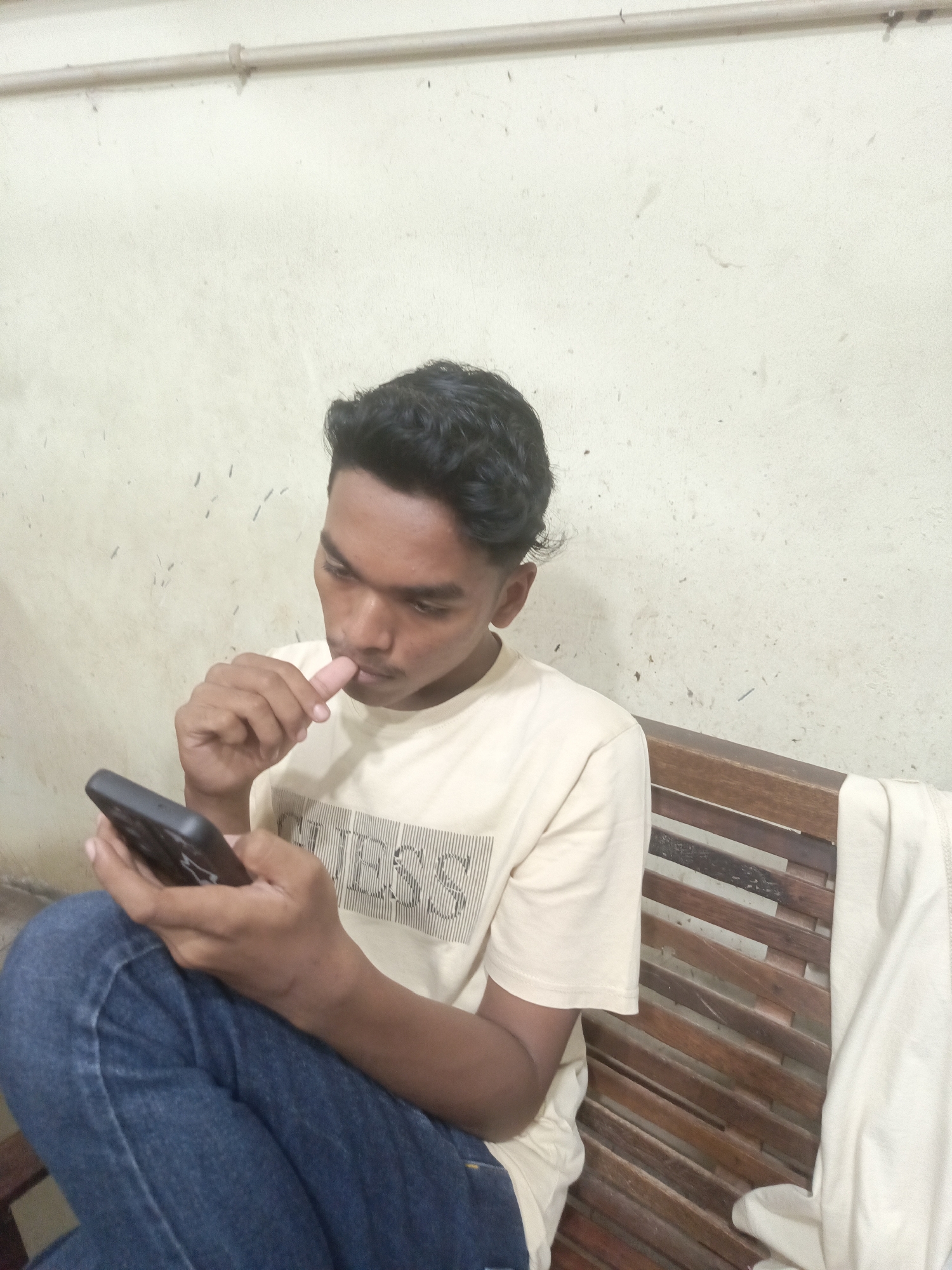 A young South Asian male, appearing to be in his late teens or early twenties, is seated on a wooden bench against a textured off-white wall. He is wearing a light-colored t-shirt with vertical black lines forming a pattern that appears to spell "GUESS" across the chest, and dark blue jeans. The individual is holding a dark-colored smartphone in his left hand and has his right index finger near his mouth, as if in thought or contemplation. A light-colored cloth is draped over the right side of the bench. The environment suggests an indoor or covered outdoor setting with basic furnishings. There is a metal pipe running horizontally along the top of the wall. The image was taken in Lhokseumawe, Indonesia.