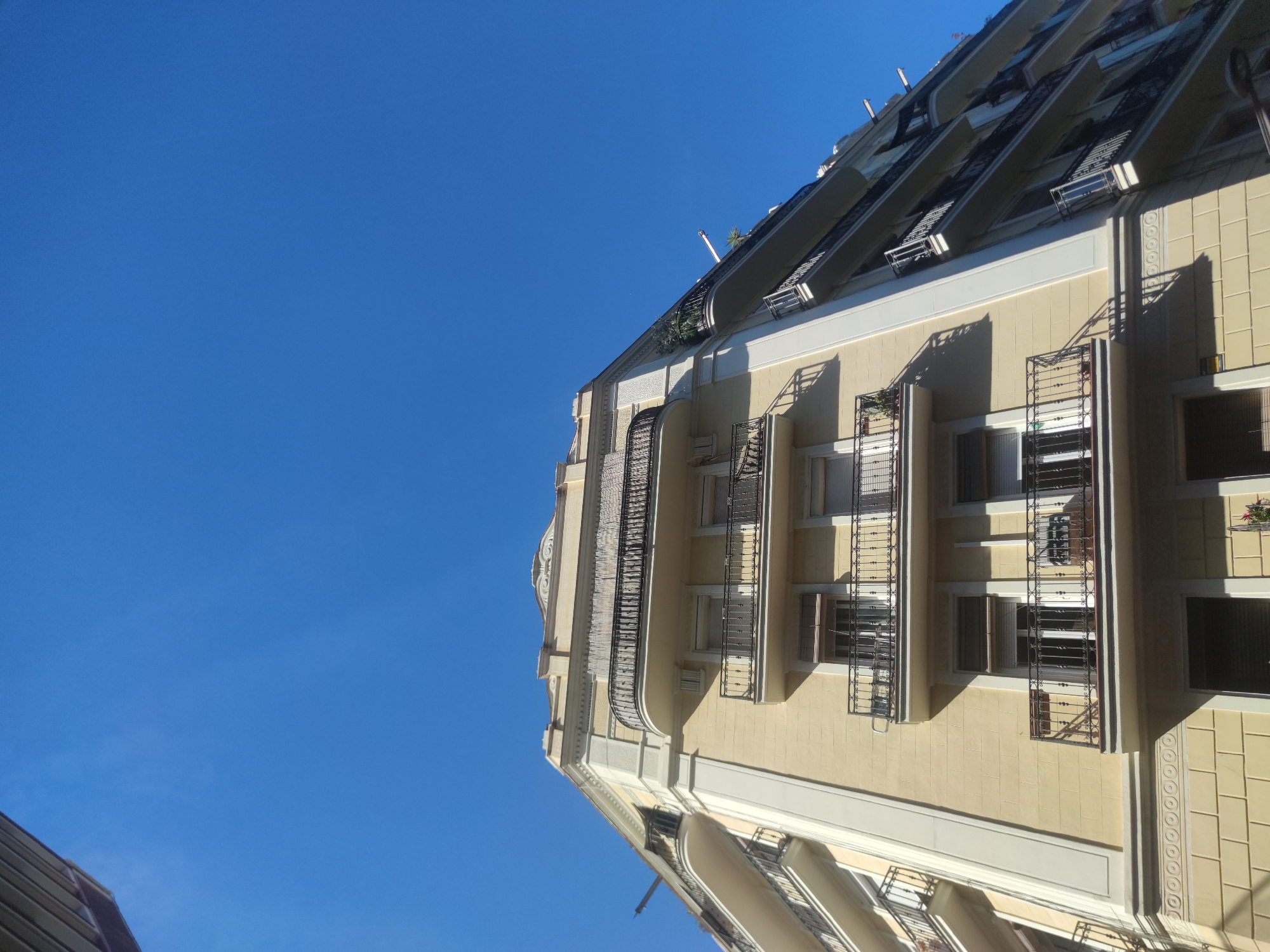 Here is a detailed description of the image:
**Scene Description:**
The image depicts a tall, light-yellow building with architectural details, including balconies with ornate iron railings and decorative moldings. The building appears to be multi-storied. The sky is a clear, bright blue with no clouds visible. The photograph is taken from a low-angle perspective.
**Objects:**
* Building
* Balconies
* Iron railings
* Windows
* Moldings
* Sky
**Environment:**
* Urban environment
* Bright, sunny day
**Location:**
* City: Unknown
* Unknown