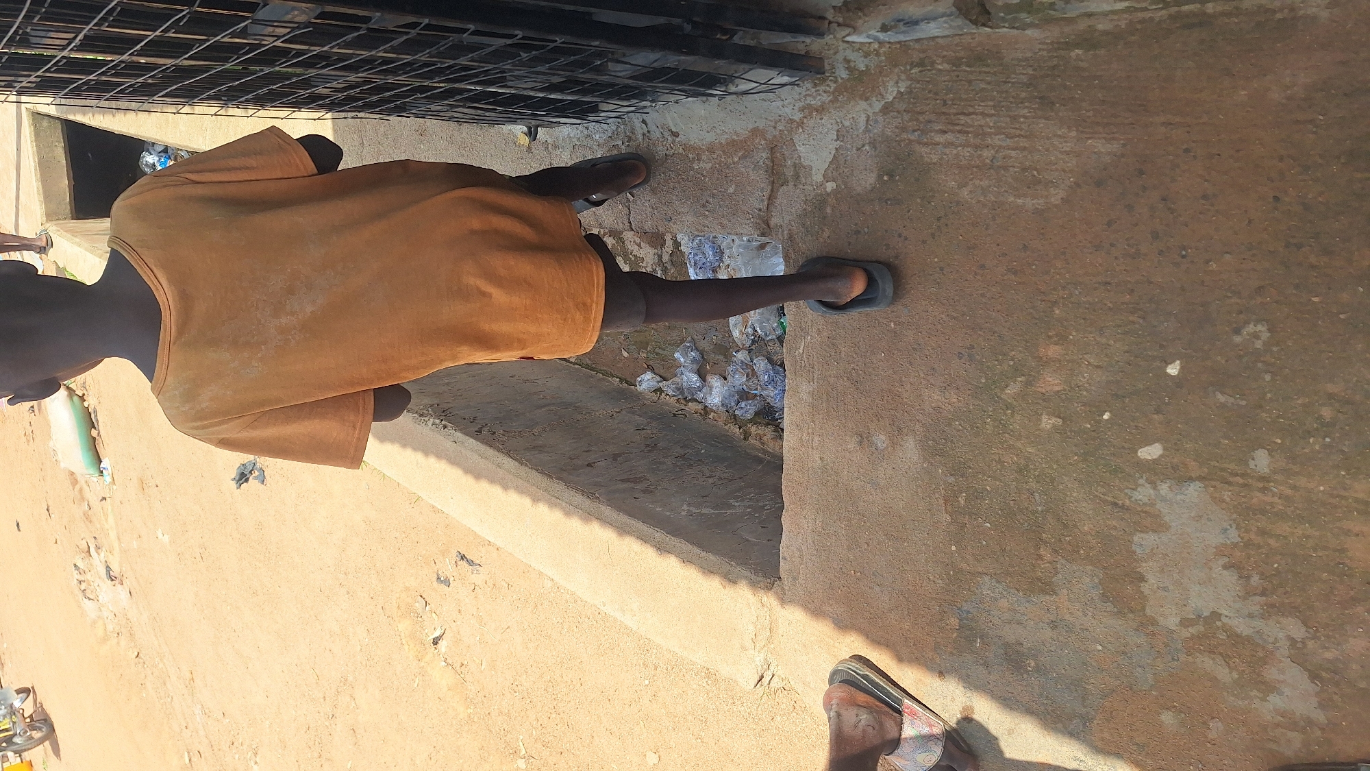 In Jalingo, Nigeria, a child, seen from behind, walks on a concrete pathway. The child wears an orange t-shirt and dark flip-flops. The pathway is adjacent to a narrow, concrete-lined drainage channel filled with plastic debris. To the left of the pathway, a dusty, unpaved area with scattered debris is visible. A metal gate runs along the right edge of the frame, partially obscuring the view of the area beyond. The overall environment suggests a neglected urban space.