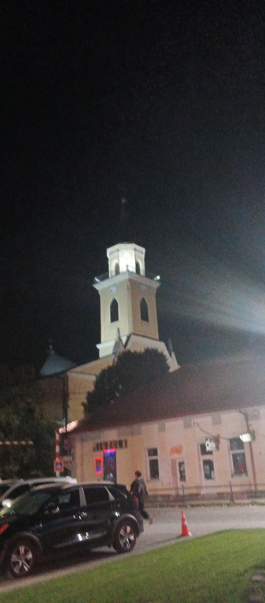 The image, taken at night in Berehove, Ukraine, shows a street scene with a prominent church tower illuminated against a dark sky. In the foreground, a dark-colored SUV is parked on the left, facing right. To the right of the SUV, a person in dark clothing is walking away from the camera. Behind them, an orange traffic cone is visible on the pavement. A beige building with several windows and signage is situated behind the cone and person, with a portion of a darker-roofed structure to the left of the building. The church tower, octagonal at the top and lighter in color, rises above the building and features arched openings. The overall impression is of a quiet urban nighttime setting, likely a street in the town center.