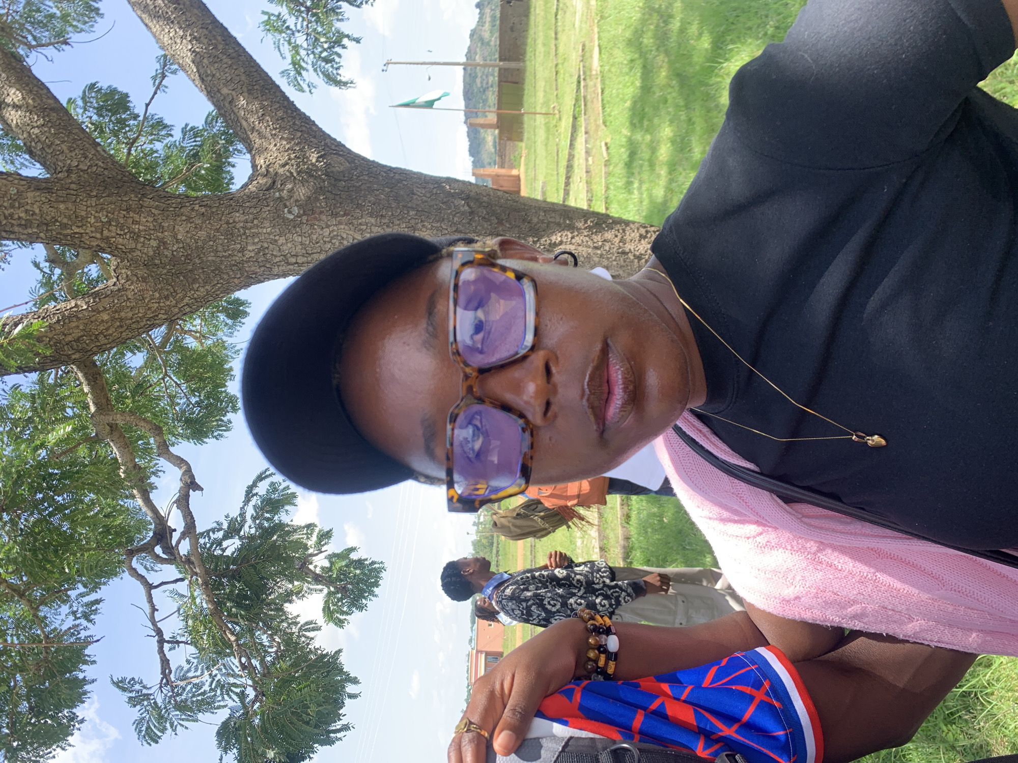 The image is a selfie taken outdoors, in what appears to be the city of Gwafan, Nigeria. The foreground is dominated by the face of a woman wearing a black t-shirt, a pink knitted cardigan, and a black baseball cap. She is also wearing large, square-framed glasses with a tortoise shell pattern and a gold necklace with a small pendant. Her left arm, visible on the left side of the frame, is adorned with several beaded bracelets and a ring on her index finger. A piece of fabric, blue with red markings, is held in her hand.
In the middle ground, several other individuals are visible. One person, a man wearing a patterned sweater and light-colored pants, is looking to the right. Another person, a woman, is wearing an orange backpack and looking to the right. Behind them, there are grassy areas, some rudimentary brick structures, and a flag pole with a green and white flag.
The background shows a tree with large branches, a clear blue sky with scattered clouds, and a hilly landscape. The overall impression is a casual outdoor gathering, possibly in a rural or semi-urban area within Gwafan, Nigeria.