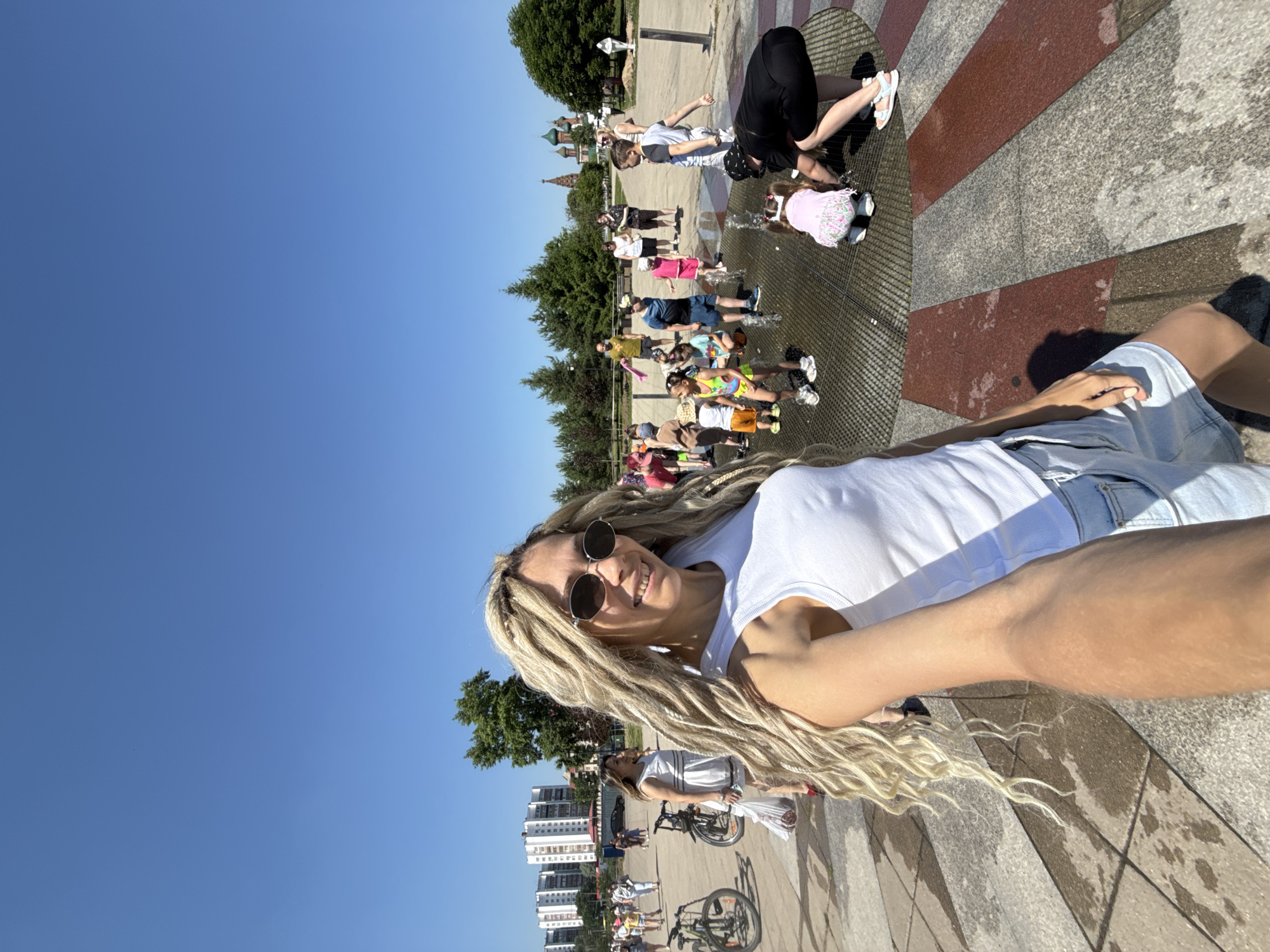 In an outdoor public space on a sunny day, a woman with blonde dreadlocks smiles at the camera. She is wearing a white tank top and denim shorts. Behind her, several children are playing in a water fountain. Other people are visible in the background, some walking and others with bicycles. The environment includes paved ground with decorative patterns, trees, and buildings in the distance. The image appears to have been taken in the city of Shebunino, Russia.