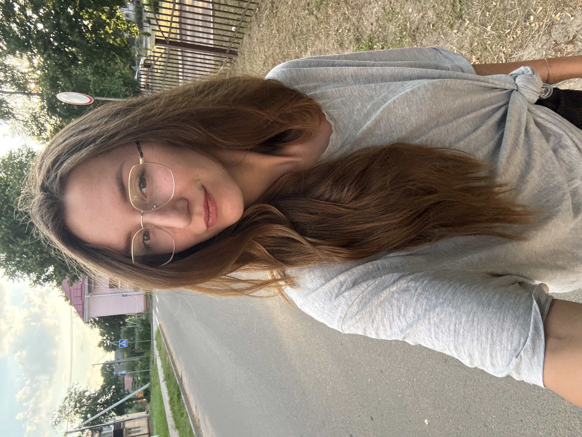 The media file is a selfie taken outdoors in Zhodzina, Belarus, with the camera held in a vertical orientation. The subject is a young woman with long, brown hair and glasses. She is wearing a light gray, short-sleeved t-shirt that is tied in a knot at her side. The background shows a paved road, a grassy area with dry vegetation, a metal fence, and trees. In the distance, a few houses and street signs are visible. The lighting suggests it is either early morning or late afternoon, with the sun casting a warm glow. The sky is partly cloudy. The woman's expression is relaxed and she is looking towards the camera. There is no visible text in the media file.