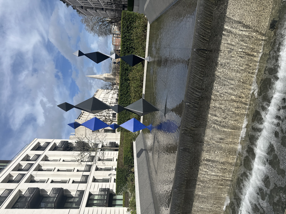 The scene depicts an outdoor urban environment within Regent's Place, London, United Kingdom, under a partly cloudy sky. In the foreground, a multi-tiered concrete fountain features water flowing over a textured vertical edge, creating white froth in a lower basin. The upper basin holds still water, reflecting the sky and sculptures.
Three abstract, faceted sculptures rise from the upper fountain basin. The leftmost sculpture is composed of three interconnected blue rhomboid-like shapes, tapering upwards. The middle and rightmost sculptures are each composed of two interconnected dark grey/black rhomboid-like shapes, resembling stylized human figures, and are reflected in the water.
Behind the fountain and sculptures, a manicured green hedge line is visible, interspersed with bare deciduous trees. In the background on the left, a multi-story, light-colored modern building with dark balconies and window frames stands prominently. A similar building is partially visible on the right. Centrally in the background, the distinctive spire of St. Marylebone Parish Church rises above the surrounding structures. No people are present.