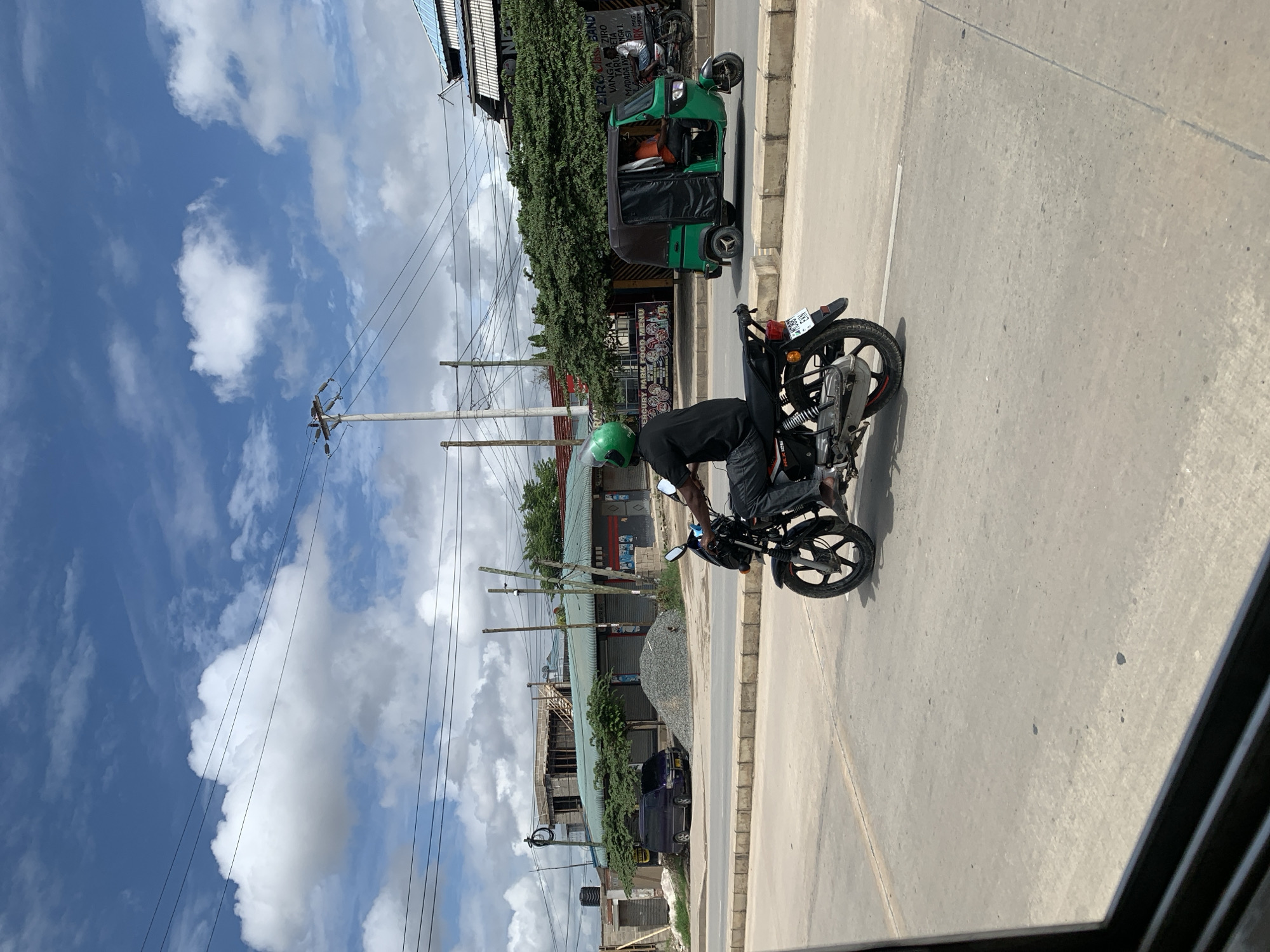 A medium-shot, sideways-oriented, eye-level photograph captures a street scene in Dar es-Salaam, Tanzania, during daylight. A person with a green helmet is on a black motorcycle. The street is paved with concrete. A green tuk-tuk is also in the scene. Power lines run along the street. Buildings and lush greenery line the street. The sky is partly cloudy.