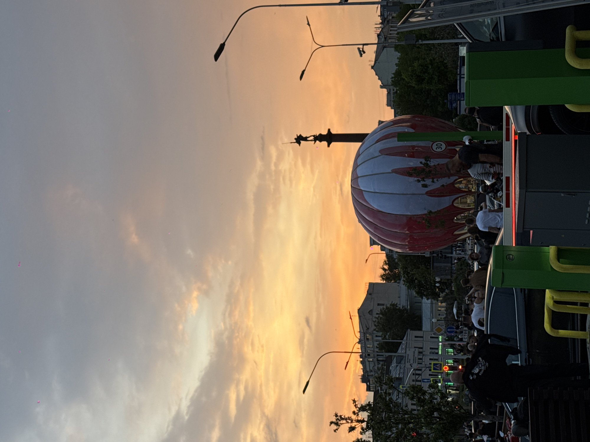 The image captures a scene in Moscow, Russia, at sunset. A large, spherical structure resembling a hot air balloon is visible, adorned with red and white stripes. Atop the structure is a monument featuring a figure. The sky is a blend of orange and gray, indicating either sunrise or sunset. In the foreground, part of an open-top bus filled with people is visible. Pedestrians can be seen near the bus. The surrounding urban landscape includes buildings and streetlights.