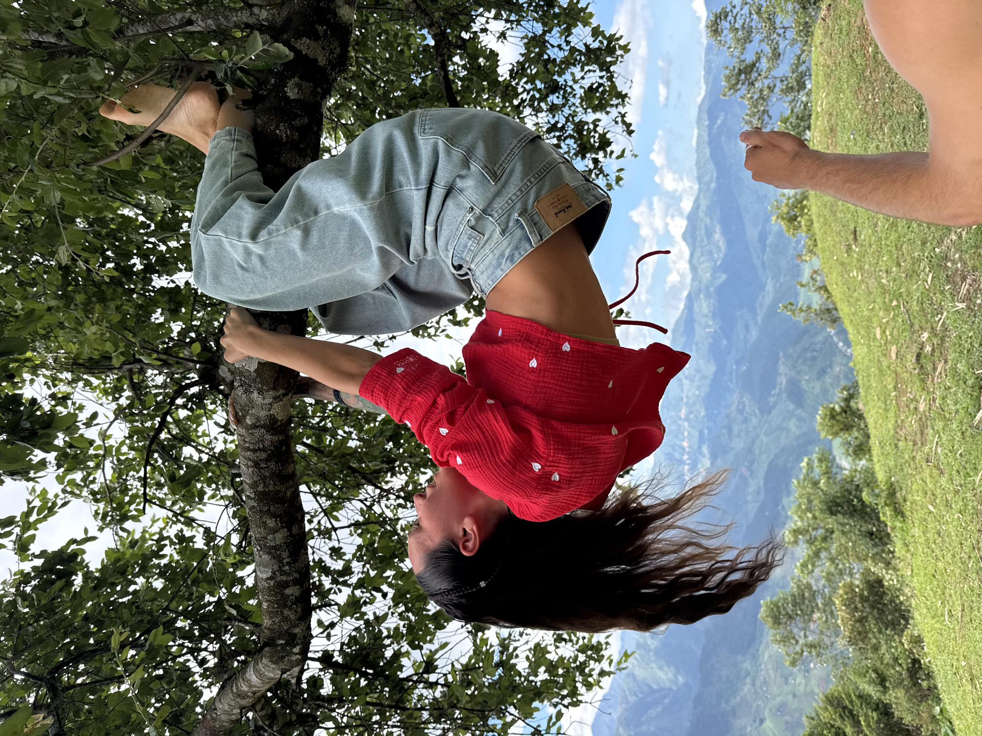 A young woman with long, dark hair is playfully hanging upside down from a tree branch, her body bent at the waist. She is wearing light blue, loose-fitting denim pants and a red, long-sleeved crop top with small white heart patterns. Her bare feet are visible, hooked over a branch to secure her position. Her left hand is gripping the tree trunk, while her right hand is not clearly visible, possibly also holding onto the tree. Her hair cascades downwards towards the ground due to gravity.
The setting appears to be outdoors, with lush green foliage surrounding her. The background reveals a scenic view of a mountainous landscape with rolling hills covered in greenery, suggesting a rural or natural environment. The sky is partly cloudy with patches of blue visible. The lighting indicates it is daytime, likely a bright and clear day. Another person's arm, with light skin and some arm hair, is visible on the far right of the image, possibly reaching out or observing. The overall impression is one of carefree enjoyment and adventure in nature. There is a small brown patch on the back of her jeans, which appears to be a brand label, but the text is not legible.