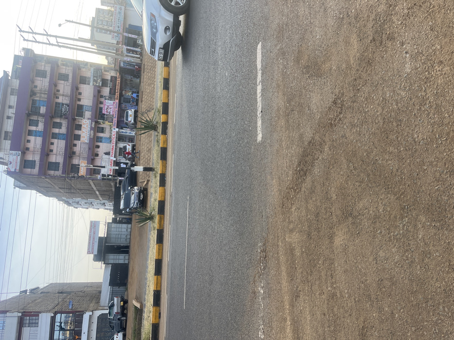 The image displays an urban street scene in Gitothua ward, Kenya, characterized by a multi-lane asphalt road, commercial buildings, and vehicles under a bright, hazy sky.
In the foreground, an unpaved dirt shoulder transitions to a two-lane asphalt road, marked by a white dashed line. A yellow and black painted concrete median separates the visible lanes from oncoming traffic or a central reservation. Beyond the median, an elevated dirt strip with sparse agave-like plants runs parallel to the road.
To the left, a multi-story building with white or light-colored facades is visible. Adjacent to it, an open lot features several grey shipping container structures, one bearing a sign for "PEATON HOMES HARDWARE." A grey pickup truck is parked within this lot.
To the right, a prominent multi-story building, approximately six to seven stories tall, dominates the background. It features a light pink/purple exterior with blue window frames or railings on its upper floors. The ground floor houses multiple businesses, including "JUMIA O" (a Jumia online pick-up point), "HAFWAK PHARMACEUTICALS," and "Shera Beauty Salon." Several vehicles, including a black SUV and a dark sedan, are parked directly in front of these establishments. Approximately five individuals are observed walking or standing on the sidewalk near these businesses.
Overhead, a network of utility wires and poles is visible against the light sky. On the far right, the front portion of a silver SUV (license plate partially visible as KCY 581U) is captured on the asphalt road, suggesting movement or recent parking.