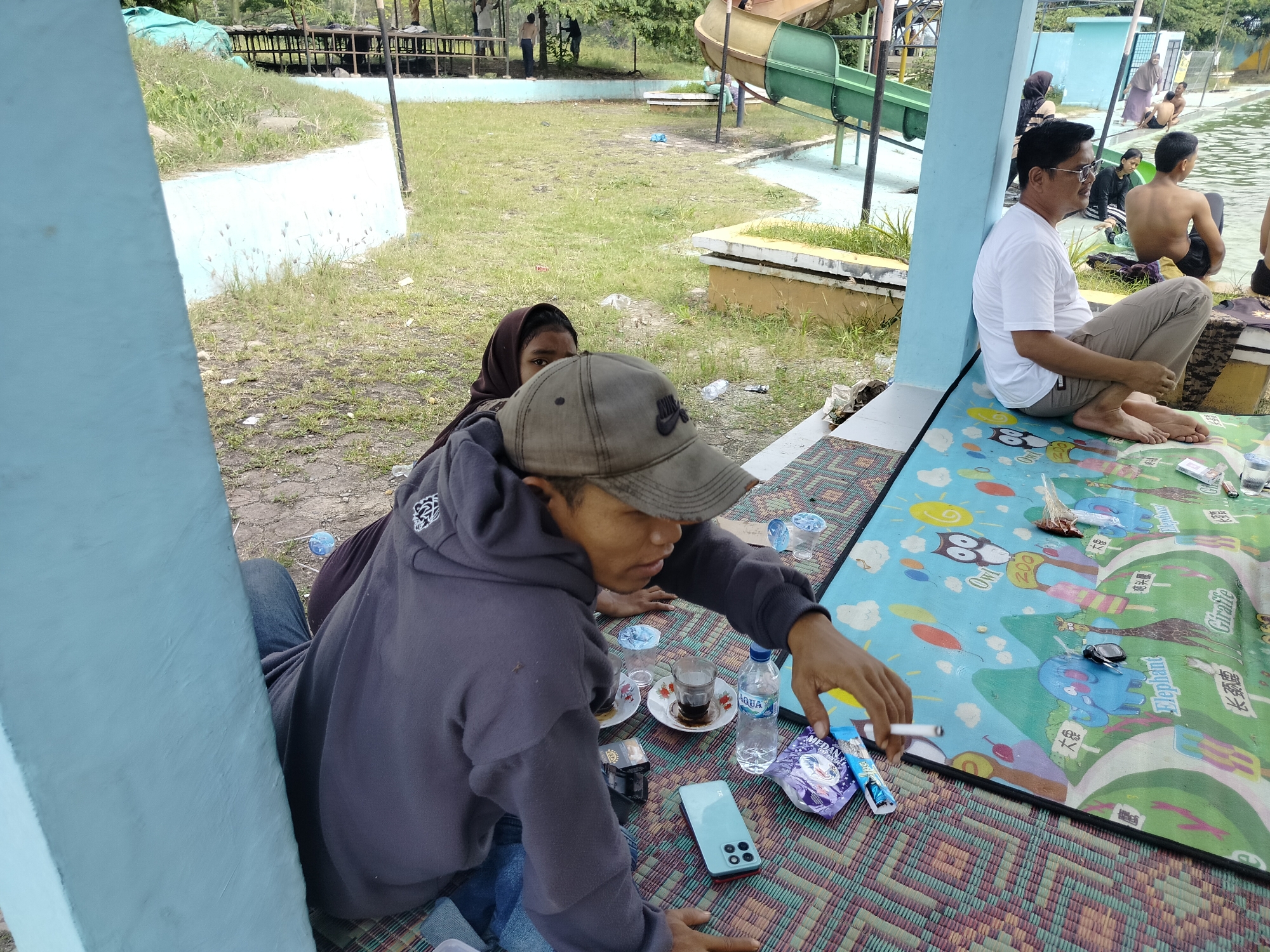 The image captures an outdoor scene at a water park or recreational area in Tegarmiko, Indonesia. In the foreground, two individuals are seated on a colorful mat. One, wearing a grey hoodie and a cap, is holding a cigarette and appears to be interacting with objects on the mat. Beside them, a child wearing a headscarf is partially visible. On the mat, there are several items including glasses of tea, bottles of water, a smartphone, and cigarette packs.
In the middle ground, a man in a white t-shirt and light-colored pants is leaning against a blue pillar. Further in the background, several other people are present, some near a swimming pool, and a water slide is visible, indicating the recreational nature of the location. The environment consists of grassy areas, concrete structures, and trees, typical of an outdoor public space.