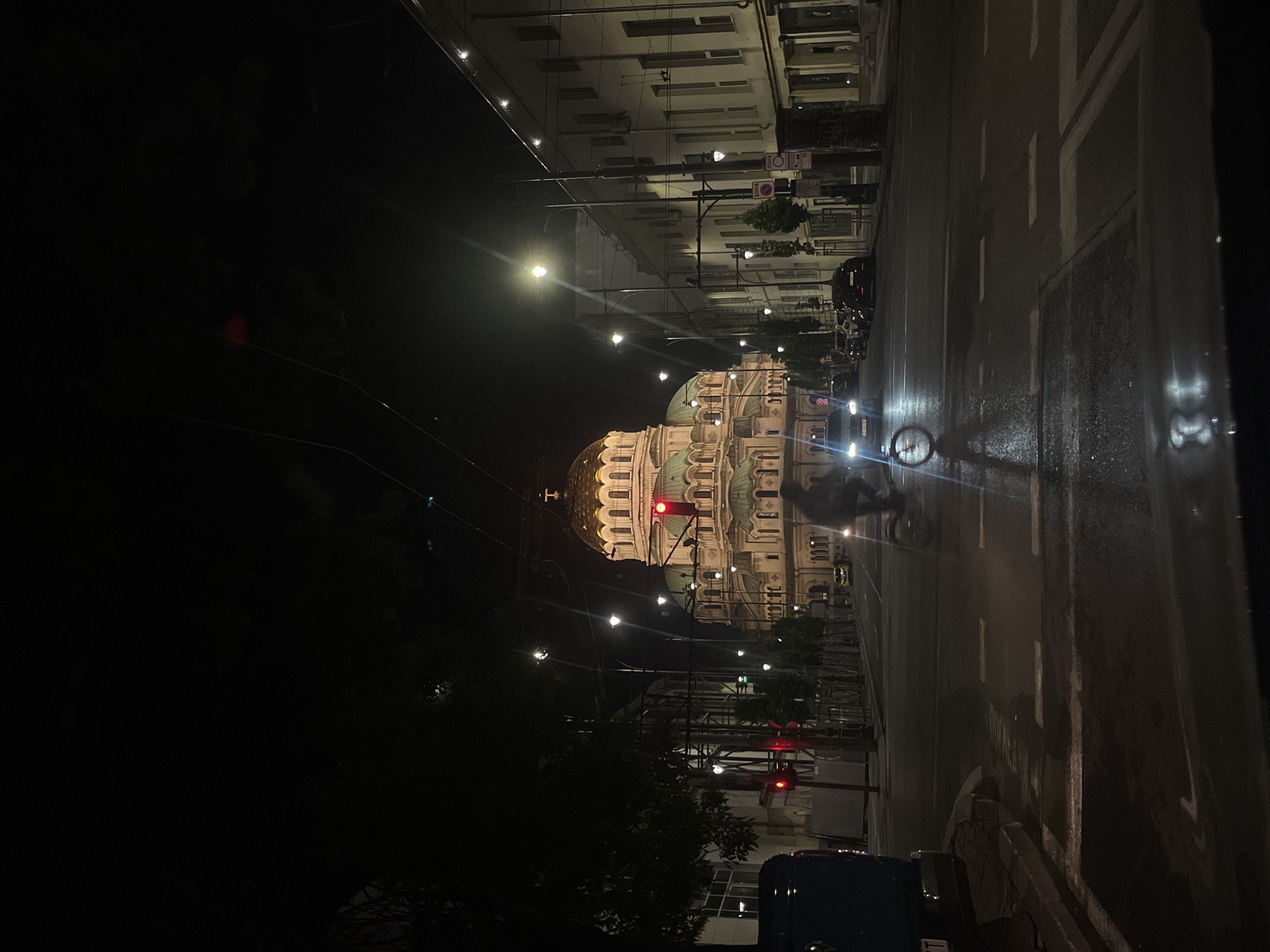 At night, the Alexander Nevsky Cathedral is seen at an angle, with a street in the city of Sofia, Bulgaria. The street is wet and slick with visible lanes. A bicyclist is seen riding towards the viewer, and there are buildings lining the street. Street lights are visible on either side of the road. There are trees, a traffic light, and a streetlight.