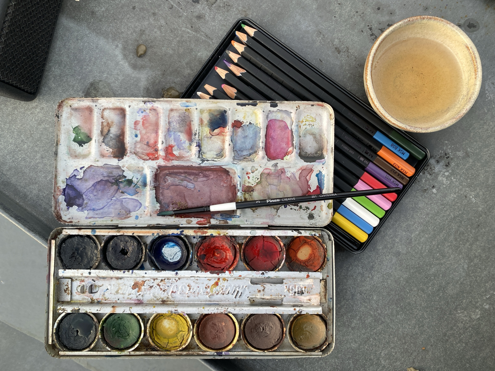 An overhead shot captures an array of well-used art supplies laid out on a textured, gray surface, likely concrete or stone, suggesting an outdoor or informal setting in Moscow, Russia.
The central items are two open metal watercolor paint palettes. The top palette features a mixing area smeared with vibrant colors like purple, red, and green, alongside several pans of heavily used watercolors. A single watercolor brush, labeled "Pinax CREATIVE 351 СИНТЕТИКА" (SYNTHETICS), rests within this palette. Below it, a larger palette displays two rows of circular, solid watercolor cakes in various states of use, including dark blues, greens, yellows, reds, and earthy browns, some showing cracks from drying.
To the right, a black plastic tray holds a set of colored pencils with black barrels and sharpened tips in a spectrum of colors. Visible text on the pencils includes "MSK B08", "PLUNGE BLUE", "MSK H01", "RAVE PURPLE", "MSK P12", "WODDHOTE BROWN", "MSK N03", and "GLOW BRANGE", with "MSK" possibly referencing Moscow.
Adjacent to the pencils, a light brown ceramic bowl holds turbid, brownish water, indicating its use for rinsing brushes. A dark, textured object is partially visible in the upper left corner. The arrangement and condition of the materials suggest an ongoing or recently completed artistic endeavor, specifically watercolor painting, under natural daytime lighting.