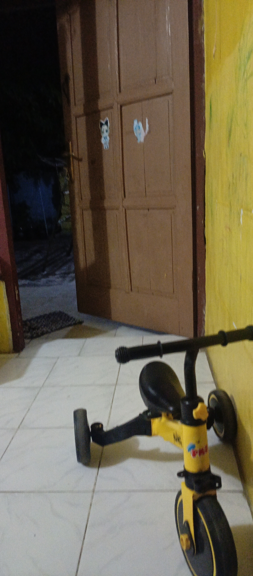 The image captures an indoor scene, possibly a hallway or entryway, with a yellow wall on the right and a brown wooden door in the background. The door has two panels and two small stickers of cartoon cats. To the left of the door, a dark opening reveals an outdoor space with some greenery. In the foreground, a yellow and black children's tricycle is positioned on a tiled floor. The tricycle has two wheels visible, one in the front and one at the back, and appears to be a balance bike or a training tricycle. The overall lighting suggests it may be evening or a dimly lit indoor space. There are no people visible in the image.