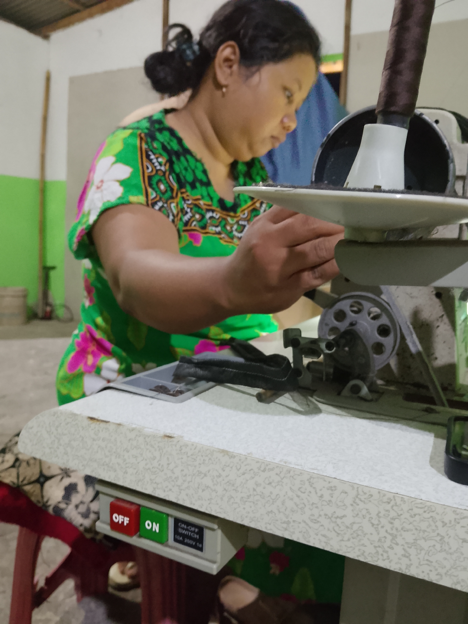 A woman with dark hair pulled back in a bun, wearing a green and white floral print dress, is focused on using a sewing machine. She is seated at a table in what appears to be a workshop or domestic setting. Her right hand is engaged with the machine's mechanism, which includes a spool of dark thread at the top. Below the table's edge, a control panel is visible with a red "OFF" button and a green "ON" button, along with a label indicating "ON-OFF SWITCH 10A 250V 1". The background shows a wall painted in muted tones, with some indistinct objects and a wooden pole. The overall environment suggests a practical workspace. This scene is likely located in Kelurahan, Indonesia, given the context of a sewing operation.
