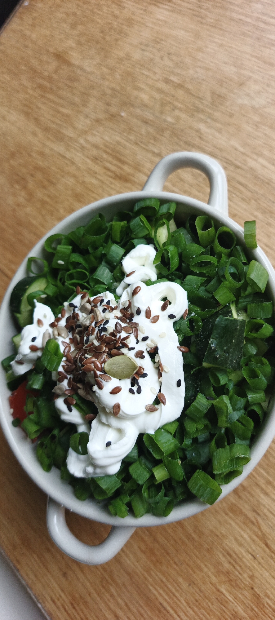 The scene is located in Chaikovsky, Russia. The image displays a light-colored, possibly off-white, ceramic bowl featuring two small handles, one visible on the upper right and another partially visible on the lower left. The bowl is filled with a prepared food item. The dominant component of the food consists of a substantial quantity of finely chopped green onions, exhibiting a vibrant green color. Several diced pieces of dark green cucumber are interspersed within the green onions, along with a small, fragmented piece of a red vegetable, possibly a tomato or bell pepper. A significant dollop of a thick, white, creamy substance, consistent with a dairy product such as sour cream or yogurt, is centered atop the green ingredients. This creamy topping is densely garnished with a mixture of various seeds. Identifiable seeds include brown flax seeds, black sesame seeds, white sesame seeds, elongated greyish sunflower seeds, and at least one distinct green pumpkin seed. The bowl is positioned on a flat, wooden surface, which displays a light-to-medium brown hue and visible wood grain patterns. No individuals, human interactions, or specific actions beyond the food's presentation are depicted.