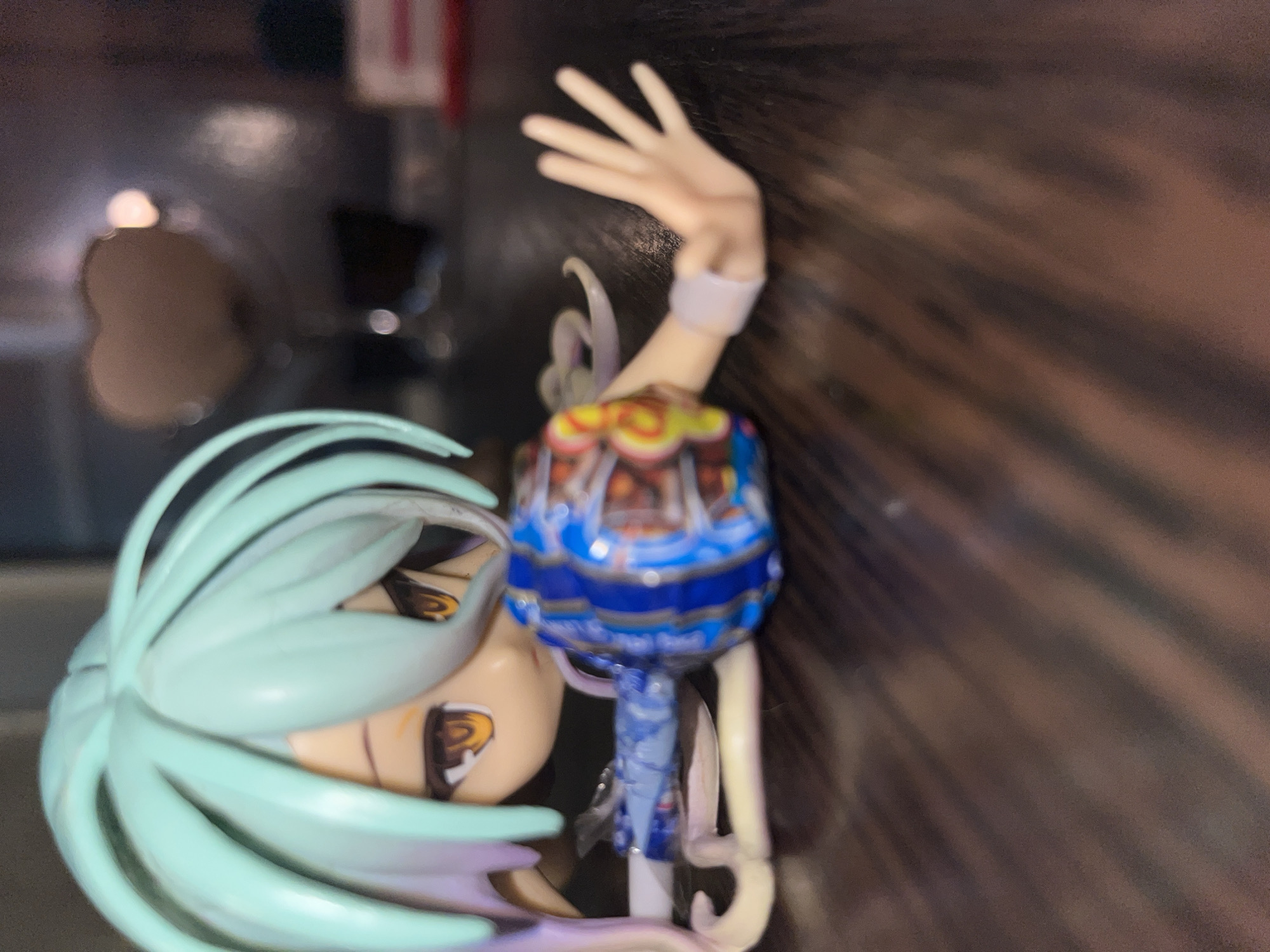 An anime figurine with light teal hair is lying on its stomach on a dark wooden surface. The figurine is wearing a blue outfit with musical notes on it and its right arm is outstretched, palm facing up. There is a colorful spherical object next to the figurine's torso. In the background, a blurred heart-shaped object is visible. The lighting suggests an indoor setting with shadows cast on the wooden surface.