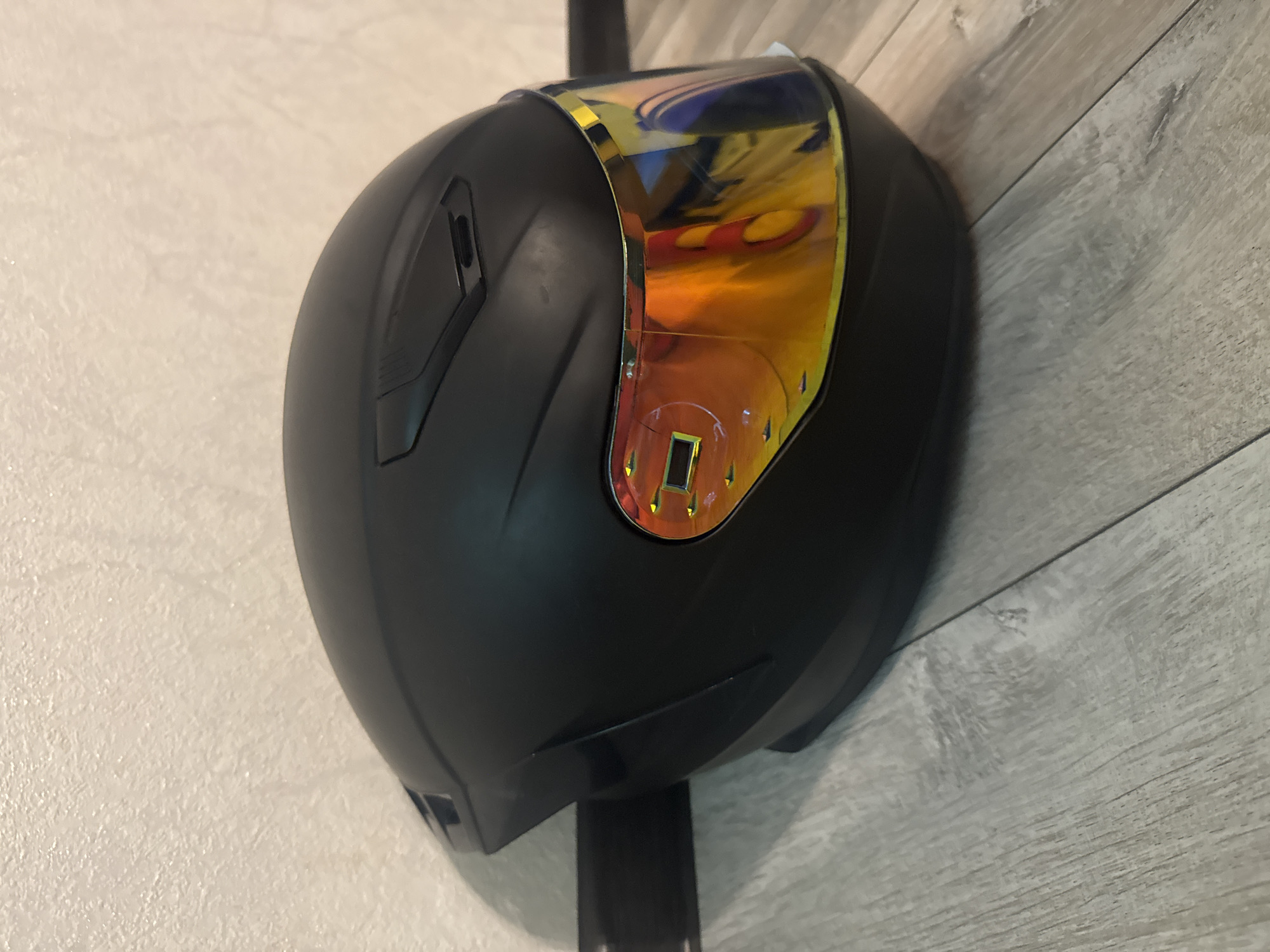 A black motorcycle helmet with a reflective orange and yellow visor is positioned on a light gray laminate floor. The helmet is partially leaning against a textured, off-white wall. A dark brown baseboard runs along the bottom of the wall. The scene is indoors and does not depict any specific locations in Bishkek City, Kyrgyzstan.