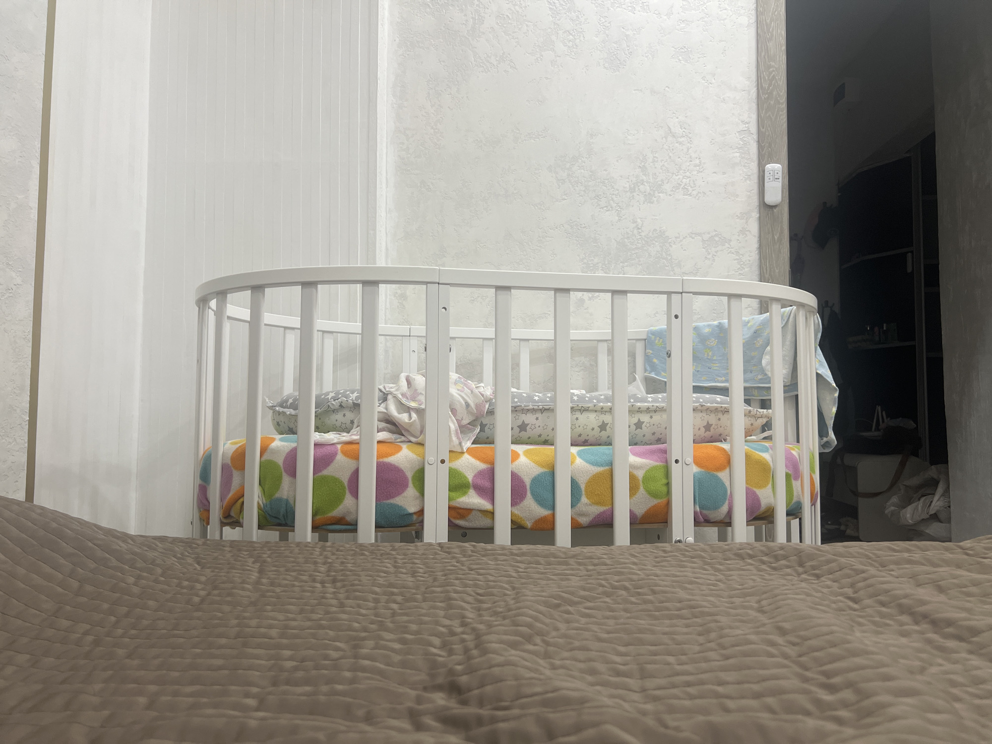 A white oval crib filled with blankets and pillows sits in the foreground of a room. The crib is made of vertical wooden slats and has a rounded top. A colorful blanket with large polka dots in green, orange, pink, and blue covers the bottom of the crib. On top of this blanket are several pillows, some with star patterns and others with floral designs. A blue and white patterned blanket is draped over the side of the crib. The crib is positioned in front of a wall with a textured, light grey finish. To the left of the crib, a vertical white paneling covers part of the wall. To the right of the crib, a narrow doorway leads into a dimly lit area where shelves and objects are visible. A white remote control is mounted on the wall to the right of the crib. The floor in front of the crib is covered by a textured brown blanket, suggesting the crib is placed on or near a bed. The room appears to be a bedroom.