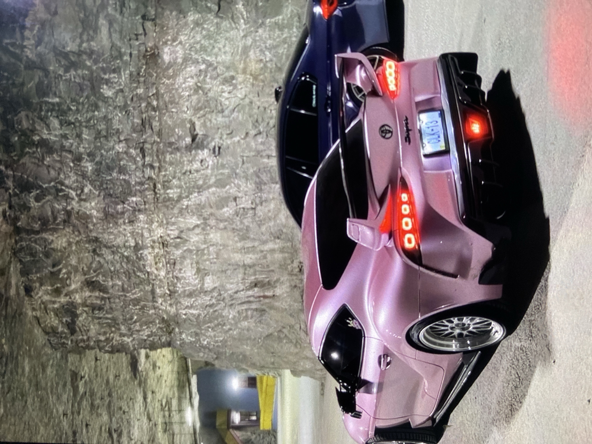 Two cars, a pink Toyota Supra and a dark blue sedan, are parked next to a stone wall. The license plate on the pink car reads "CLK-13." The photo was taken in a quarry or tunnel-like structure. There is no evidence to confirm the location is Lhokseumawe, Indonesia.