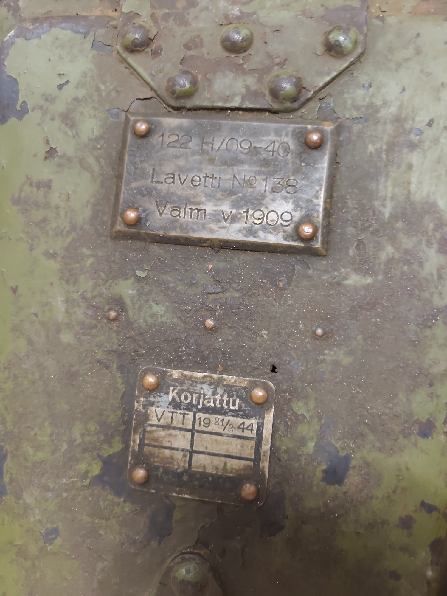 The image shows a close-up of a weathered green metal surface with two rectangular metal plates riveted to it. The top plate has embossed text that reads "122 H/09-40 Lavetti No 138 Valm. v 1909". The bottom plate has embossed text that reads "Korjattu VTT 19 21/2 44". The metal surface is chipped and shows signs of rust. The lighting is diffused, and the focus is on the metal plates. The background is out of focus. This image is not located in the city of Viimsi, Estonia.