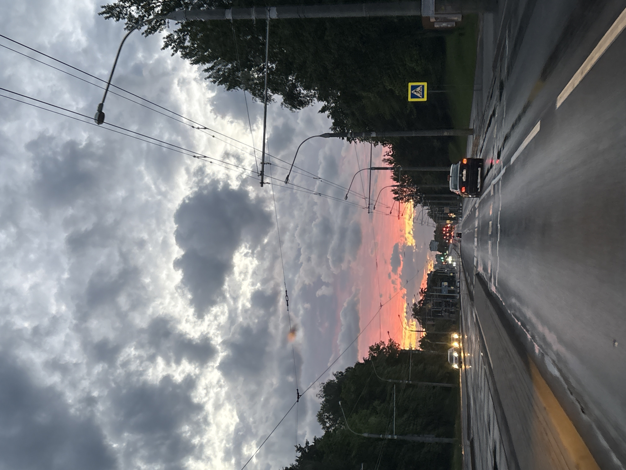The image shows a street in Minsk, Belarus, under a dramatic sky with a sunset visible between buildings and trees. The road surface is wet, reflecting light from the sky and streetlights. A car is driving away in the center lane, and other vehicles are visible further down the road. Overhead tram lines and lampposts extend across the street. A yellow and black pedestrian crossing sign is on the right. Trees line both sides of the street.