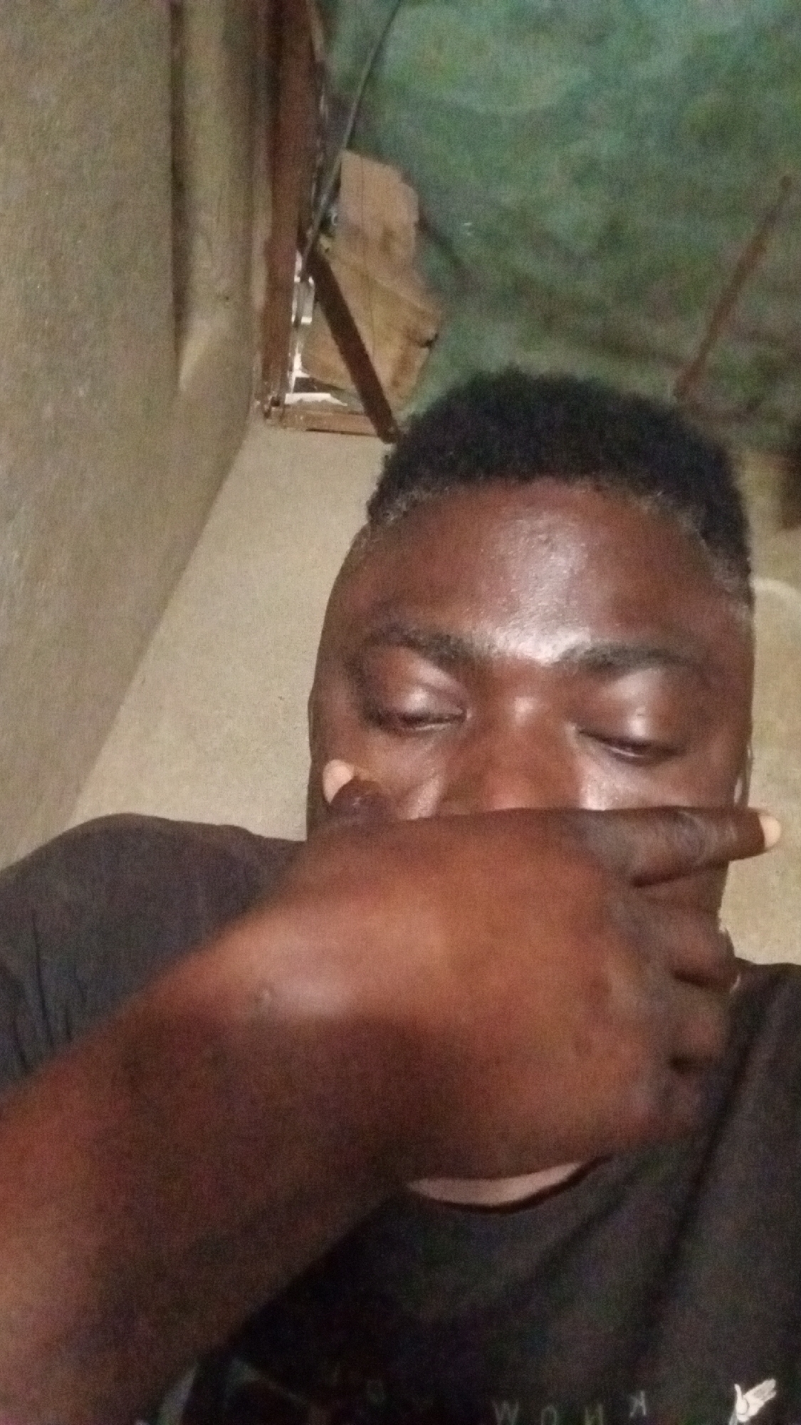 A close-up portrait shows an individual with dark skin and short, dark, textured hair, captured from a slightly low angle. The person is wearing a dark-colored t-shirt. Their right hand is raised, covering their mouth and nose, with fingers extended horizontally across the face. The eyes are closed or cast downwards, and the forehead appears shiny.
The background suggests an indoor setting. To the left, there is a light-colored, possibly concrete or plaster wall. To the right and top, the background is less defined, showing what looks like wooden planks or beams, and a green, possibly fabric or tarp-like material. These elements suggest a rustic, unfinished, or makeshift interior. The lighting is dim and diffused.
On the bottom edge of the image, partially visible on the person's shirt, the letters "W O H K" can be seen, possibly part of a larger word or logo, appearing somewhat distorted or upside down. The person's expression and pose, with the hand covering the face and closed eyes, could convey introspection, shyness, or weariness.