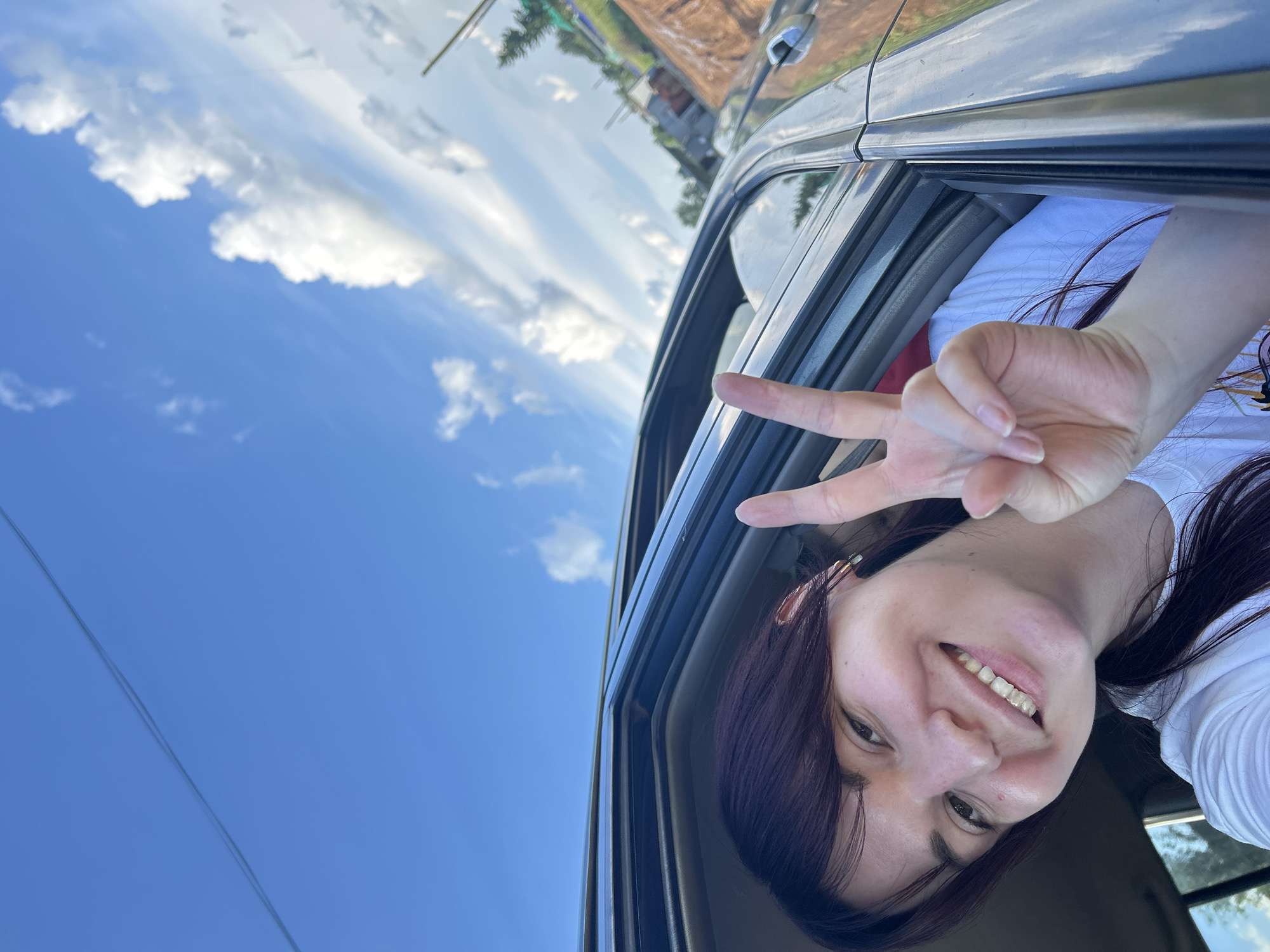 In the city of Fokino, Russia, a young woman with dark hair and bangs, wearing a white t-shirt, poses for a selfie from inside a car with her arm and head leaning out the window. She is smiling and holding up two fingers in a peace sign. The sky above is a bright blue with scattered white clouds. A power line is visible stretching across the top of the frame. The background shows a rural or semi-urban landscape with buildings and what appears to be an unpaved road or construction area. The sunlight suggests it is daytime.