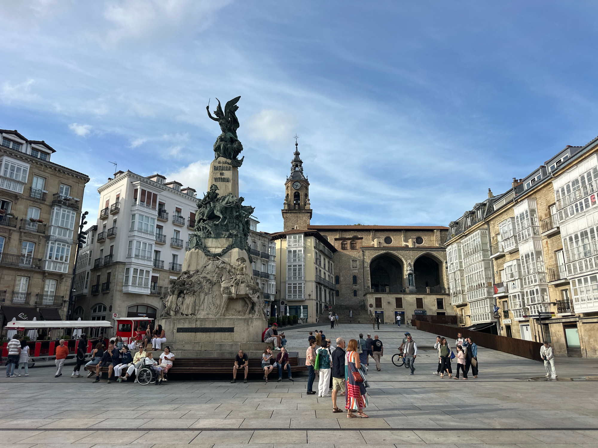 The image depicts a public square in Vitoria-Gasteiz, Spain, specifically Plaza de la Virgen Blanca, a well-known landmark. Dominating the center of the square is the Monument to the Battle of Vitoria, a tall stone pedestal topped with bronze sculptures, including a winged victory figure and figures on horseback. Surrounding the square are multi-story buildings with traditional architecture, characterized by balconies with white shutters and ornate facades.
In the foreground, a paved plaza is populated by several individuals. Some are seated on benches, including a woman in a wheelchair. Others are standing or walking, engaged in various activities such as conversation, strolling, or operating a bicycle. A red and white tourist train is visible on the left side of the square. In the background, a church with a prominent bell tower stands against a clear blue sky with scattered clouds. The overall scene suggests a sunny day with moderate public activity in a historic urban setting.