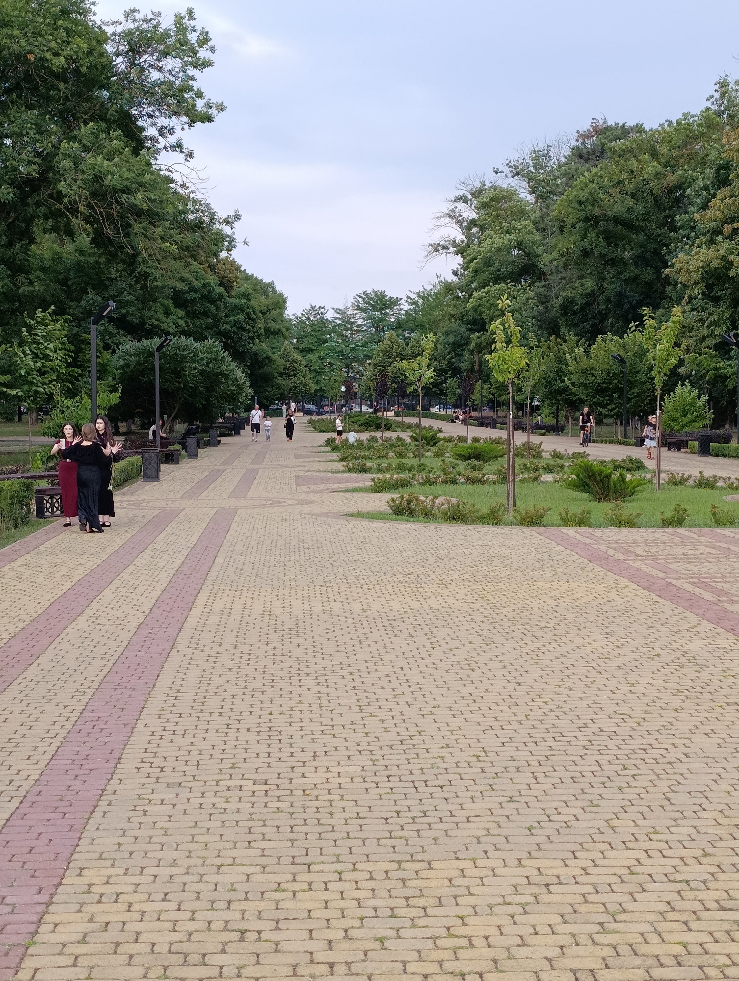The image depicts a paved park pathway with a pattern of light yellow and pink bricks, lined with green manicured bushes and young trees. In the background, a dense line of mature trees creates a lush green backdrop under a cloudy sky. Several individuals are present in the park. On the left, three young women in formal attire are standing and interacting. Further down the path, other people can be seen walking, including a man on a bicycle and a couple. The overall setting suggests a public park within the city of Maykop, Russia, likely during daylight hours with overcast weather.