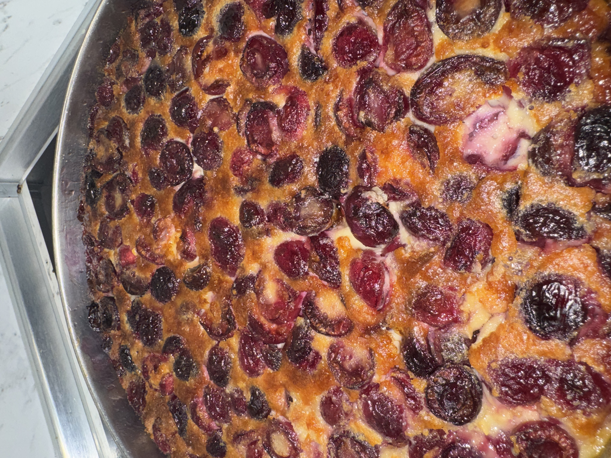 The image shows a close-up view of a cherry clafoutis in a metal baking pan. The clafoutis is richly studded with dark red and black cherries, baked into a custard-like batter that is golden-brown in places. The dessert appears to be freshly baked and is sitting on a metal surface, likely a kitchen counter or serving area. The location is Elhovo, Bulgaria.