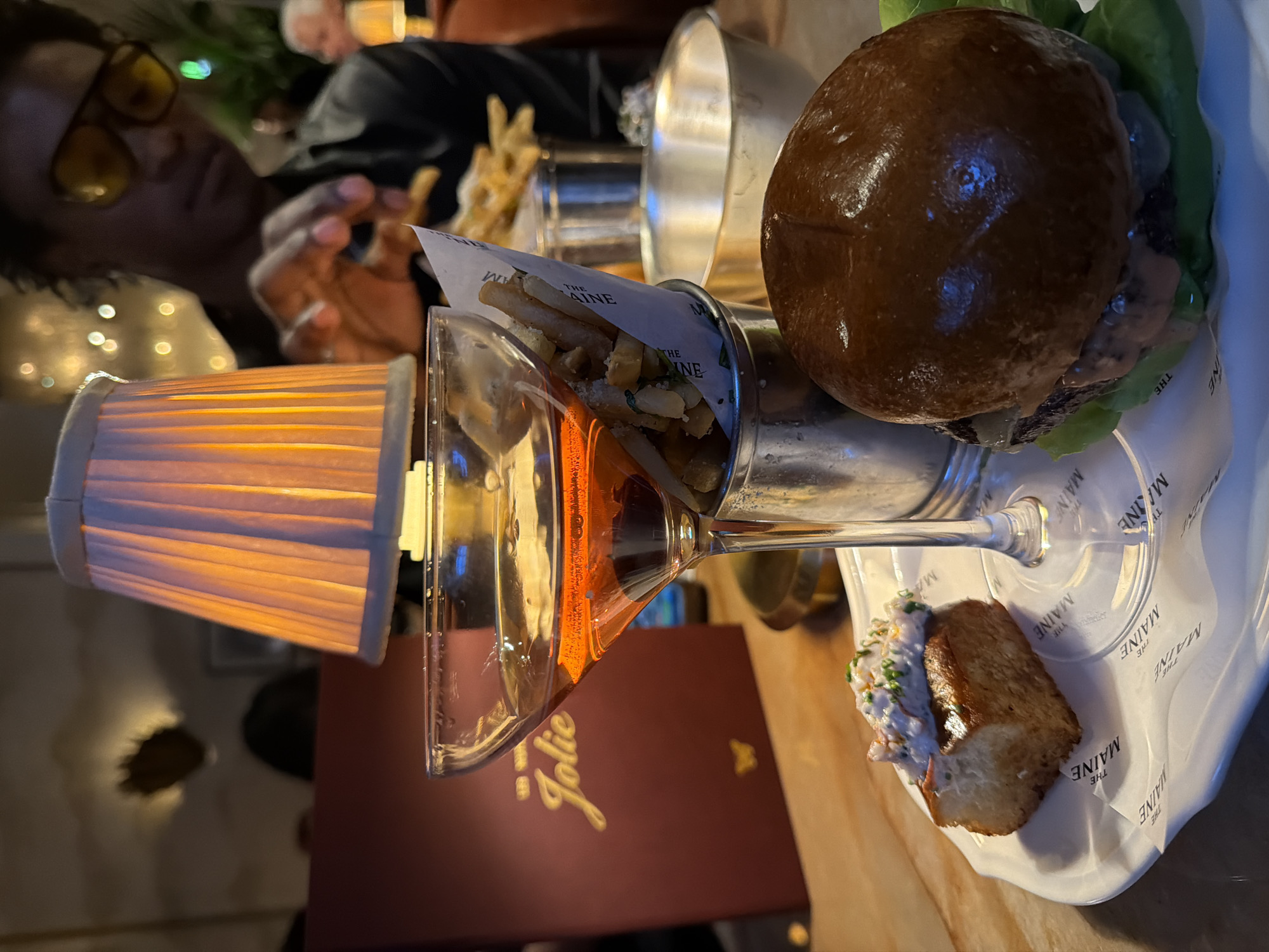The image depicts a warmly lit dining scene at the Jolie Oyster & Cocktail Bar within The Maine Mayfair, located in the city of London, United Kingdom.
In the foreground, a white, irregularly shaped plate rests on a wooden table, covered by branded paper featuring "THE MAINE" logo repeatedly. On the left side of the plate, a small, golden-brown piece of toast or brioche is topped with a creamy white mixture, possibly a seafood salad, garnished with finely chopped green herbs and what appear to be small orange specks. Dominating the center, a stemmed coupe glass contains an amber-hued cocktail. Adjacent to the glass, a small, silver metal bucket holds french fries, some visibly seasoned with green herbs and white specks (likely cheese or salt). The fries bucket is partially wrapped in paper also bearing "THE MAINE" logo. To the right, a mini burger with a dark, glossy bun, a visible patty, green lettuce, and a pinkish sauce is presented on the same branded paper.
In the mid-ground, an illuminated table lamp with a pleated fabric shade casts a warm orange glow across the scene. Behind the lamp, a person with dark, curly hair and yellow-tinted glasses is partially visible, appearing to reach for the fries with their hands. They are wearing a dark long-sleeved garment. A dark red menu or book with "Jolie" written in gold script and a small gold butterfly emblem is visible to the left, behind the cocktail glass.
The background is softly blurred, showing a dim restaurant interior. Distant bokeh lights suggest decorative lighting. A decorative wall feature, possibly a mirror or artwork with a spiky or leaf-like gold frame, reflects light on the left. The general ambiance is intimate and cozy, characteristic of an upscale dining establishment. Another person, an older individual with white hair, is faintly visible in the far right background.