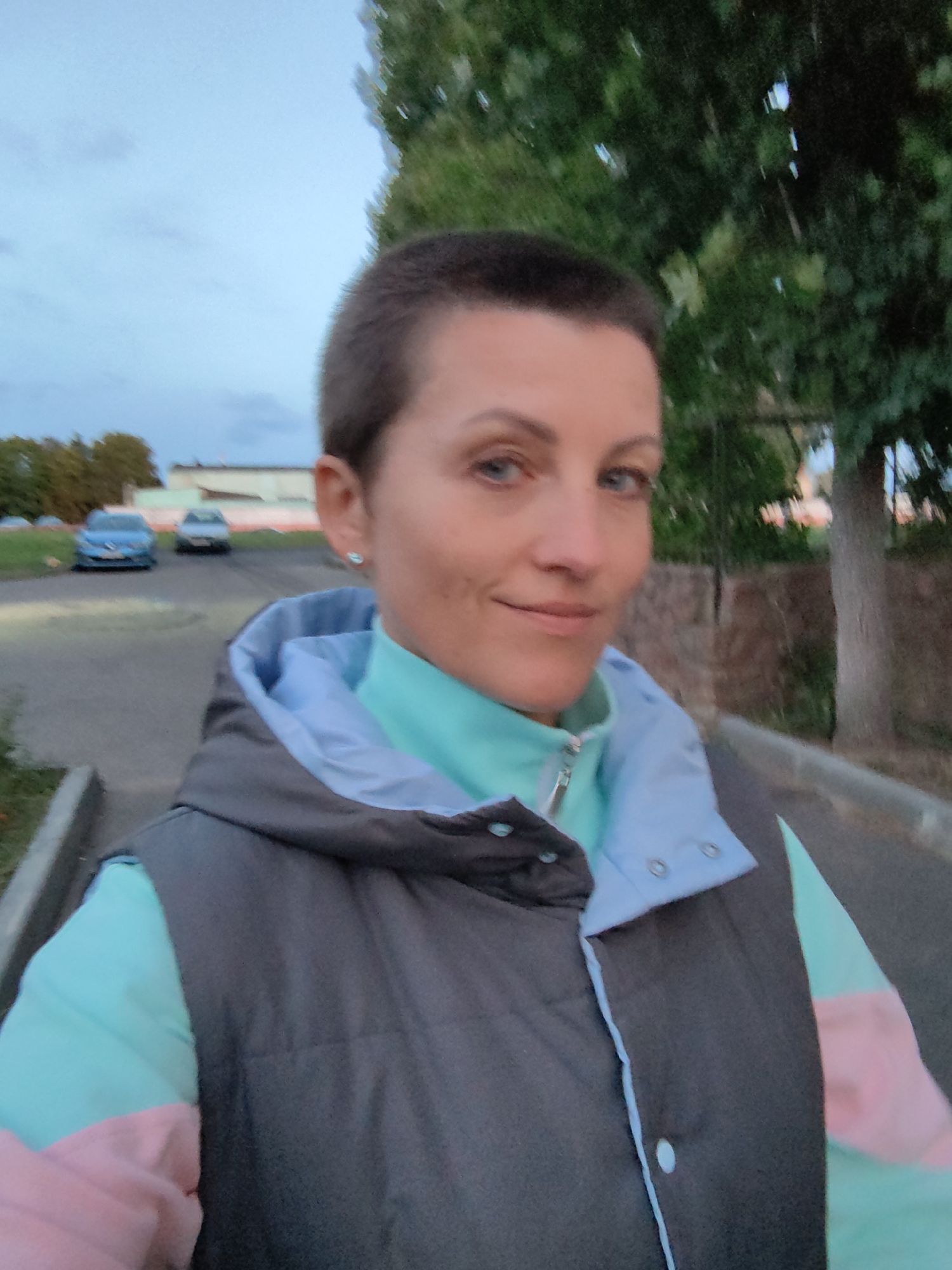 A woman with a buzzed haircut is pictured in an outdoor, urban setting, likely in Myadzyel, Belarus. She is wearing a grey puffer vest over a teal and pink jacket. Her expression is neutral, and she is looking towards the camera. The background shows a paved path, green trees, and two parked cars, one blue and one silver. The sky is overcast, suggesting it might be late afternoon or early evening.