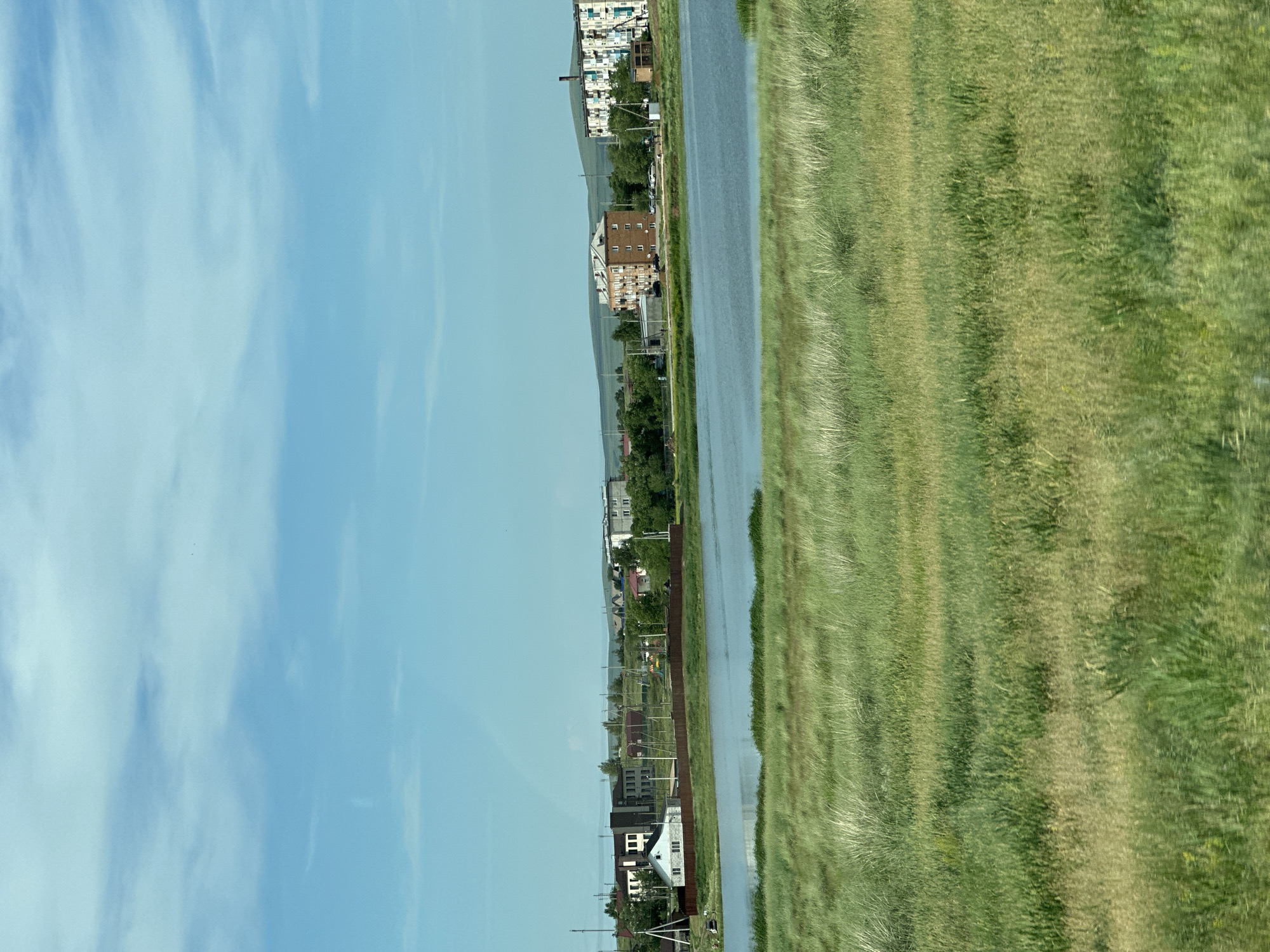 The image shows a wide shot of a settlement by a body of water, likely in the city of Борец, Russia. In the foreground, a field of tall, green grass extends towards the water's edge. The water itself appears calm and reflects the blue sky above, which is dotted with sparse white clouds.
In the middle ground, a settlement is visible on the far bank of the water. Several buildings of varying sizes and architectural styles are present. To the left, there are smaller, possibly residential houses, some with white siding and dark roofs. Further to the right, there are taller, multi-story apartment buildings with light-colored facades and balconies. Some trees are scattered throughout the settlement, and a brown wooden fence runs along the perimeter of some properties. There are also utility poles and wires visible.
In the background, rolling hills can be seen, covered in vegetation. The overall scene depicts a rural or semi-rural environment with a mix of residential and possibly some commercial or public buildings. The lighting suggests it is daytime, likely a clear or partly cloudy day. No people or specific actions are discernible in the image.
