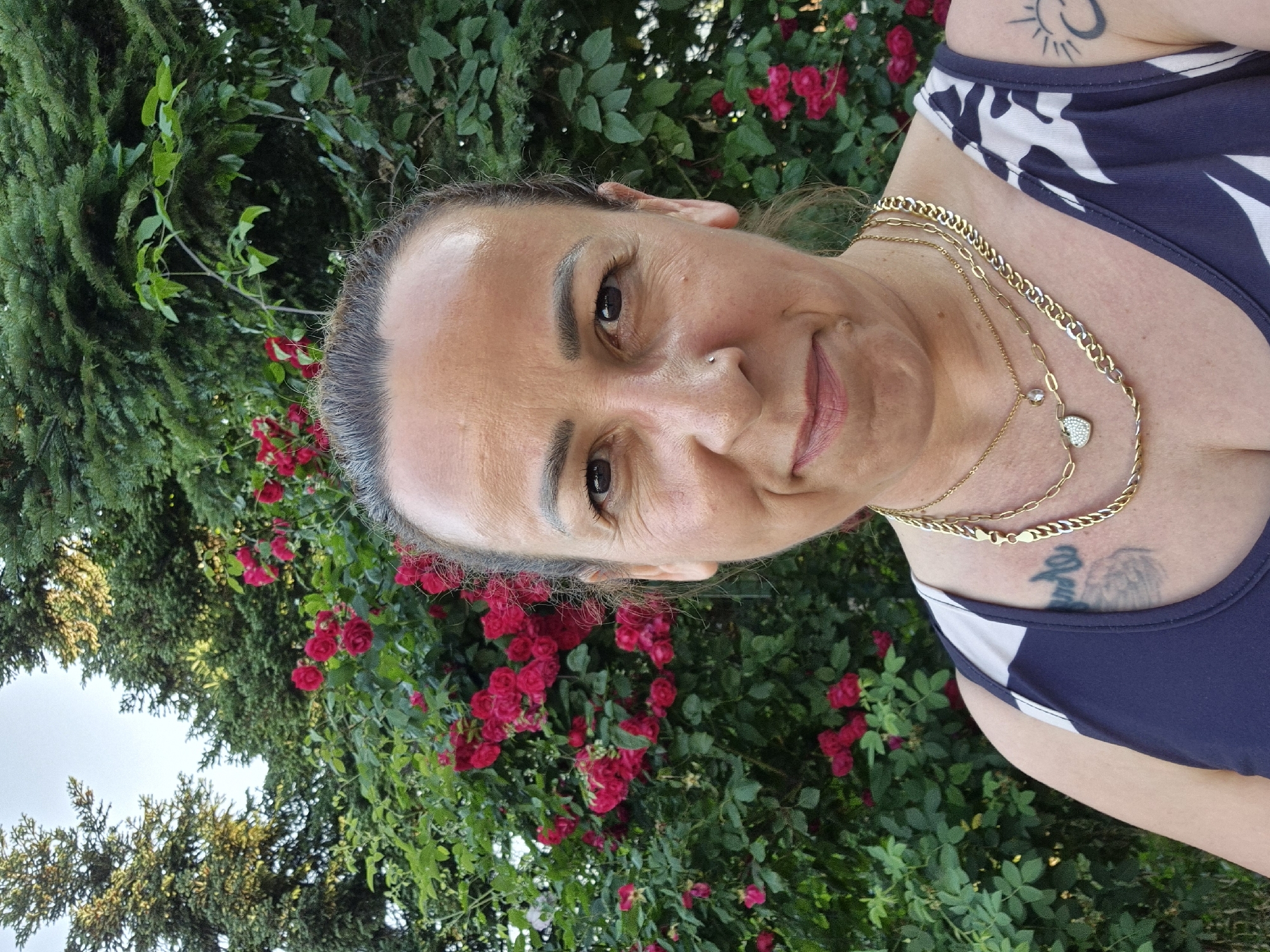An outdoor close-up portrait shows a woman looking directly at the viewer with a slight, pleasant expression. She appears to be middle-aged, with her hair pulled back, revealing some grey strands at her temples. A stud piercing adorns her nose. She wears multiple layered gold-toned necklaces; one features a small, heart-shaped pendant with what appears to be clear stones, and another has a clear, faceted teardrop bead.
She is dressed in a dark blue sleeveless top with white patterns. Two tattoos are visible: a black sun-like symbol on her upper left arm/shoulder, and another indistinct, dark tattoo partially visible on her chest area.
The background is rich with green foliage. Dense evergreen trees are visible on the left, while abundant bushes with clusters of bright red roses are prominent behind her and to her right. The scene is illuminated by natural daylight, suggesting it is daytime in a garden or outdoor setting.
