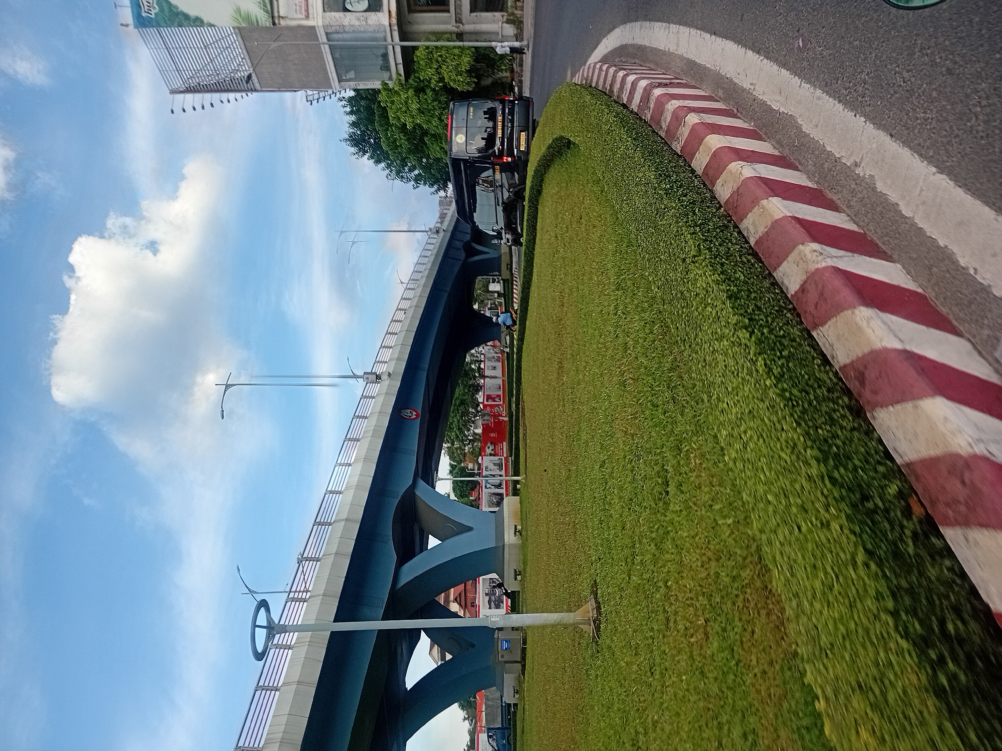 The image captures a scene in the city of Đà Nẵng, Vietnam, focusing on an elevated blue highway overpass that spans across the frame. In the foreground, a meticulously manicured green lawn bordered by a low, dense hedge curves along the roadside. The road itself features a concrete curb painted with alternating red and white stripes, typical of traffic safety markings. A black minivan, identified by the license plate "39B 06615", is prominently positioned in the middle ground, moving away from the camera on the right side of the road. A lone individual is visible on the sidewalk to the right of the minivan. Beneath the overpass, a vibrant red wall with white signage can be seen, suggesting a commercial area or public space. The sky above is a clear blue, dotted with fluffy white clouds, and a streetlamp with a circular fixture stands sentinel on the left side of the frame. The overall atmosphere is that of an urban environment with functional infrastructure and well-maintained public spaces.