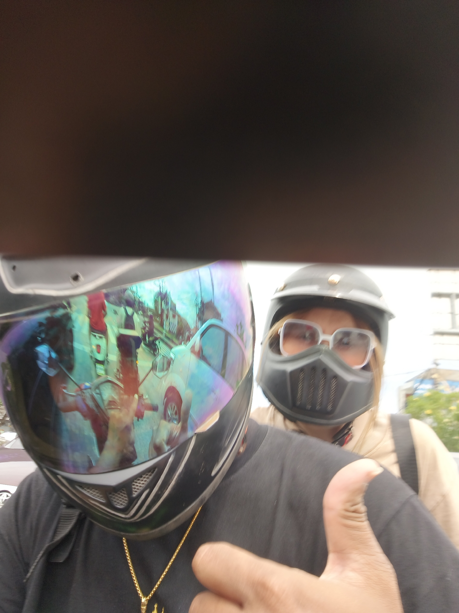A close-up selfie shows two individuals on a motorcycle, their faces obscured by helmets and a face mask. The person in the foreground, on the left, wears a black helmet with a reflective visor that captures a distorted reflection of the surrounding street scene. The reflection shows other vehicles, buildings, and people in a busy urban environment, likely Denpasar, Indonesia. The person in the background, on the right, wears a black helmet, a black face mask covering their lower face, and large, light-colored sunglasses. Both individuals appear to be wearing casual clothing. The selfie is taken from a low angle, with the upper part of the frame dominated by a dark, blurry area, possibly the edge of a roof or structure. A hand with a thumb raised is visible in the lower right corner, indicating the person taking the photo.