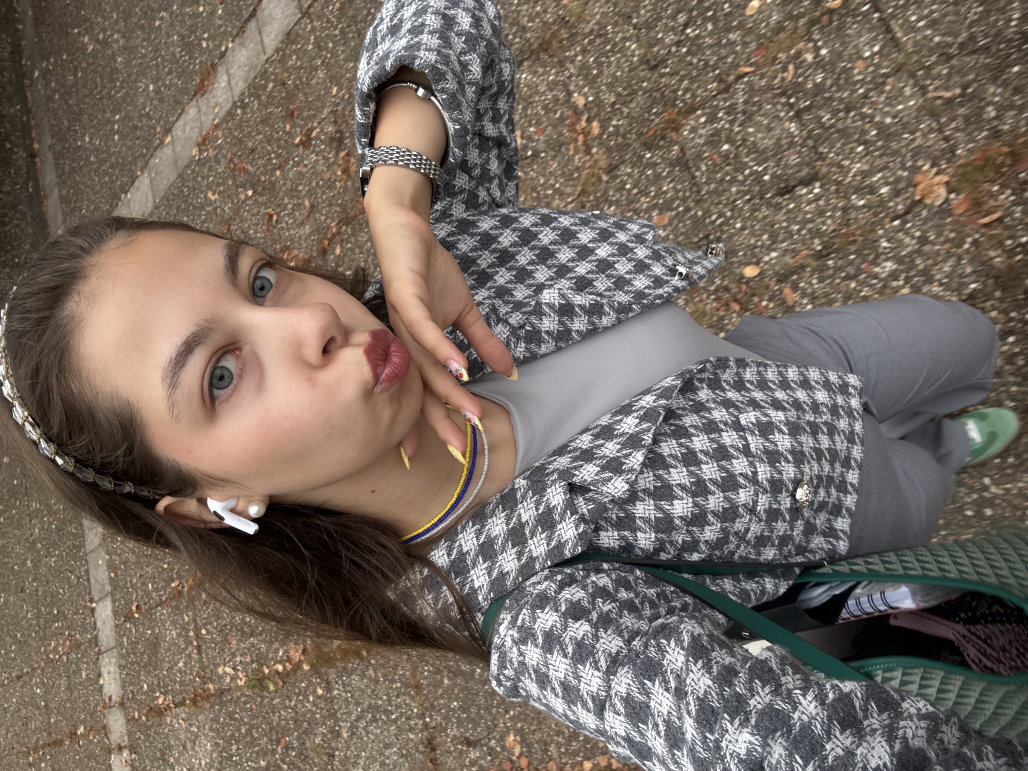 This is a portrait-oriented, slightly tilted selfie of a young woman taken outdoors. She is wearing a grey and white houndstooth jacket over a grey top and grey pants. She has blue eyes, brown hair, and is wearing a silver headband, a silver bracelet, and white earbuds. A colorful beaded necklace with yellow, blue, and grey beads is visible around her neck. Her right hand is near her face, with fingernails painted yellow and decorated with small designs. She is puckering her lips as if for a kiss.
The background is a textured grey paved surface with scattered brown leaves, suggesting an outdoor setting like a park or sidewalk. A green bag with a diamond pattern is visible near her hip, with the contents partially visible, including a notebook. The lighting suggests it is daytime. The overall mood is casual and playful. There is no visible text in the image. The provided location context indicates this scene is in Hanau, Germany.