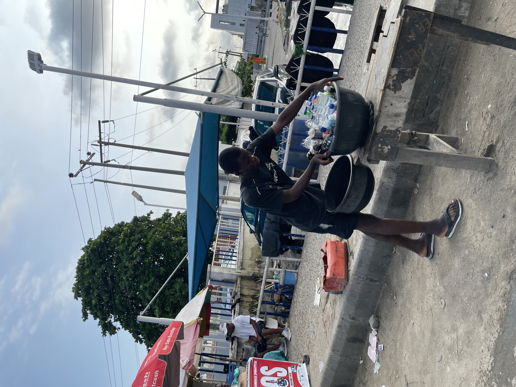 In Lekki, Nigeria, a young man is seen on a sidewalk, sorting through a collection of plastic water bottles. He is wearing a black t-shirt and dark shorts, with sandals on his feet. He is leaning over a large black basin filled with the bottles, seemingly separating them. Beside him, another black basin rests on a weathered wooden table. In the background, several cars are parked, and a red jerrycan is visible on the curb. The scene is set against a backdrop of overhead power lines and a partially visible building with a modern architectural style, suggesting an urban environment. The sky is partly cloudy.