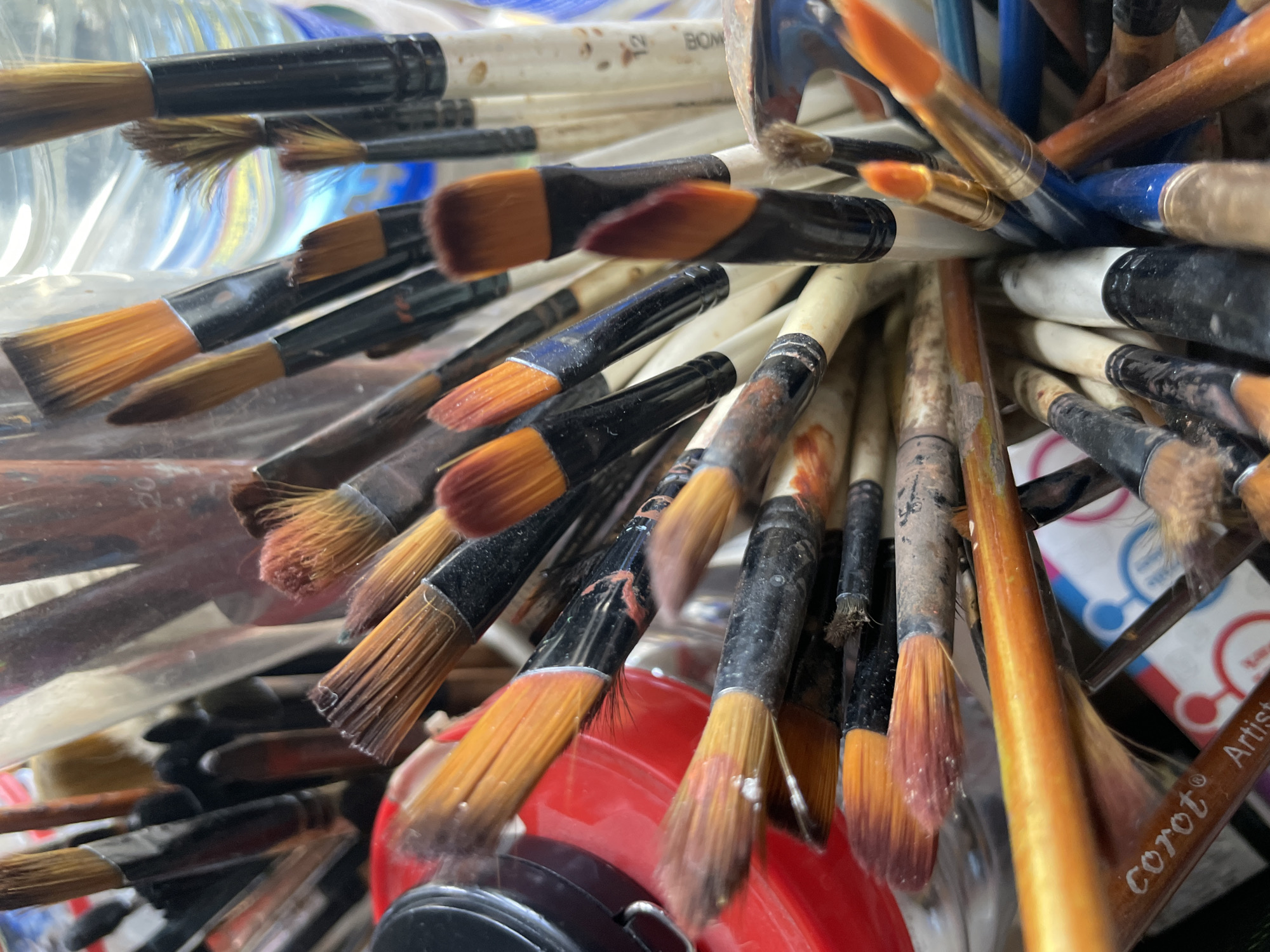 A close-up photograph depicts a collection of used paintbrushes, densely packed and presumably stored in a clear container. The bristles display a variety of colors, predominantly brown and orange, reflecting previous use with different paints. The handles vary in color and material, with some being wood and others plastic. Paint residue is visible on both the bristles and handles of several brushes. The background includes a glimpse of a red container and a white surface with what appears to be a logo or design that includes the word "Artist". Given the context of Ado, Nigeria, it is presumed to be located there. No people are visible. The setting suggests an artist's studio or workspace.