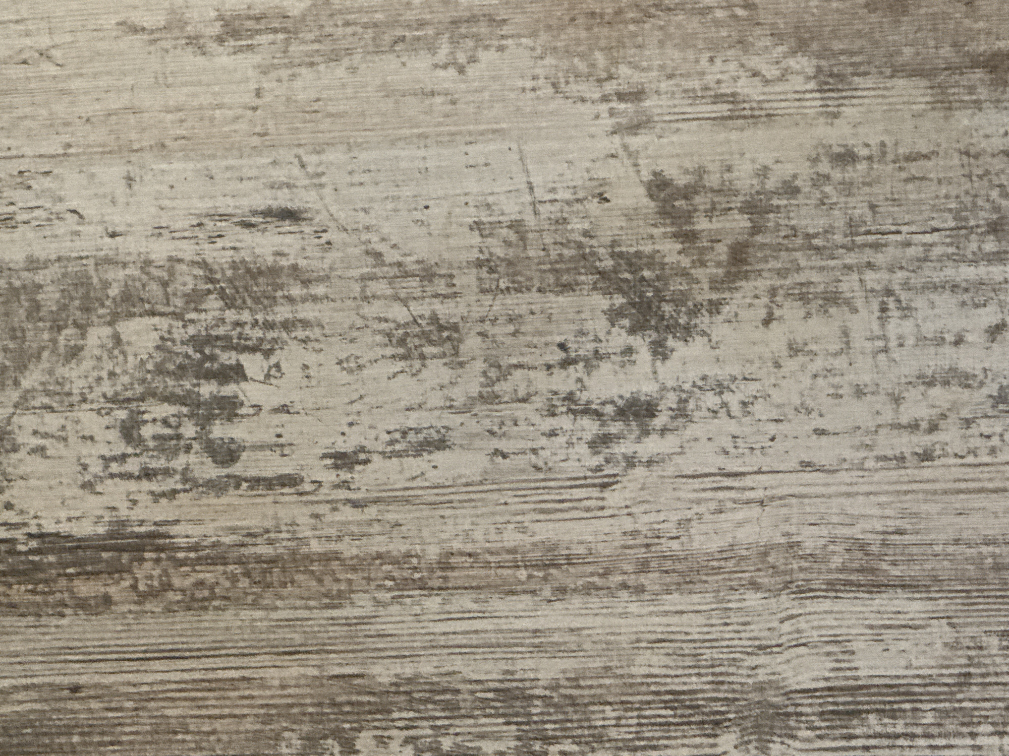 This is a close-up, overhead shot of a textured surface. The surface appears to be made of wood or a wood-like material, with a distressed or weathered finish. The color palette is muted, consisting of shades of light gray, beige, and dark brown. Horizontal lines, characteristic of wood grain, are prominent and vary in thickness and intensity, suggesting a rustic or aged appearance. There are darker, irregular patches and streaks scattered across the surface, giving it a mottled or stained look. Some faint, thin scratch marks are also visible, further contributing to the worn aesthetic. The lighting is even and diffused, with no strong shadows, indicating it might be an indoor setting or an overcast day. The image is abstract in nature, focusing solely on the texture and pattern of the material, and does not depict any people, objects, or a specific scene. No text is visible in the image.