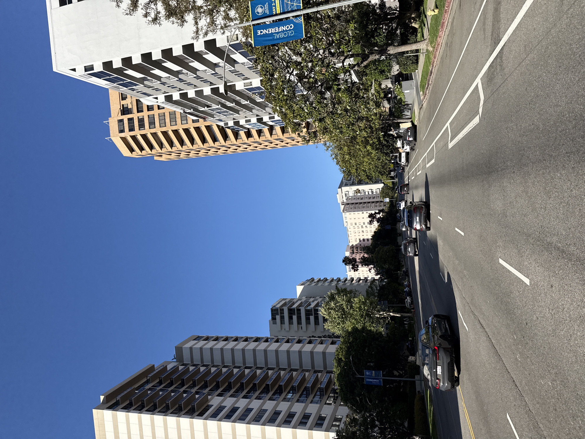 The image depicts a street scene in Los Angeles, United States of America, under a clear blue sky. Tall, modern high-rise buildings line either side of the street. The buildings are predominantly white and tan, with repeating patterns of windows and balconies. A road runs through the middle of the frame, with cars traveling in both directions. There are painted lane dividers, including lines and arrows. Green trees line the sides of the road, interspersed with street signs and light poles. A banner is visible on one of the poles, showing "Global Conference".
