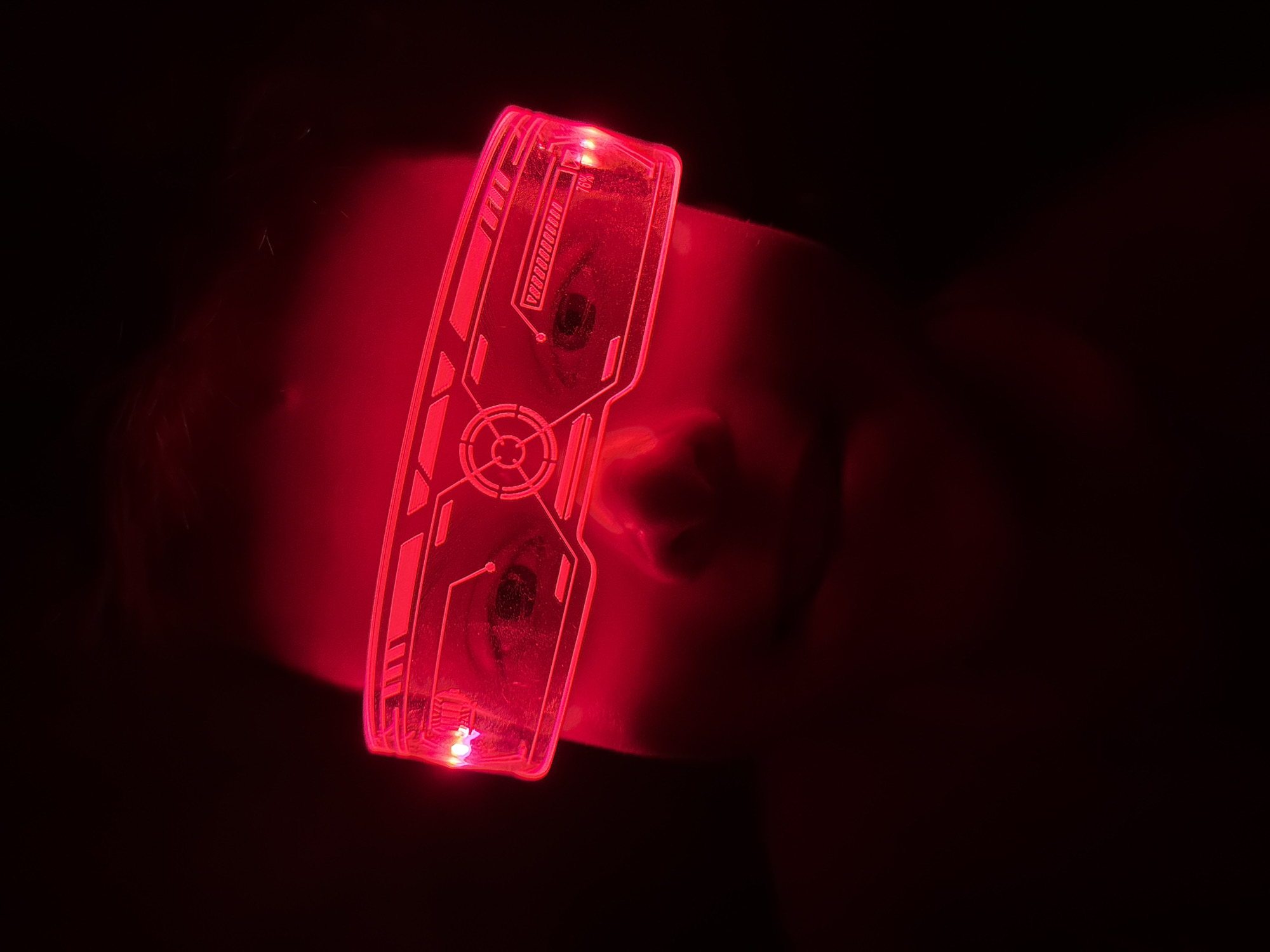 A person's face is visible in near darkness, illuminated by red light emanating from a pair of futuristic, glowing red glasses they are wearing. The glasses obscure the person's eyes, but the nose and mouth are visible. The environment is dark. There is no indication of a location.