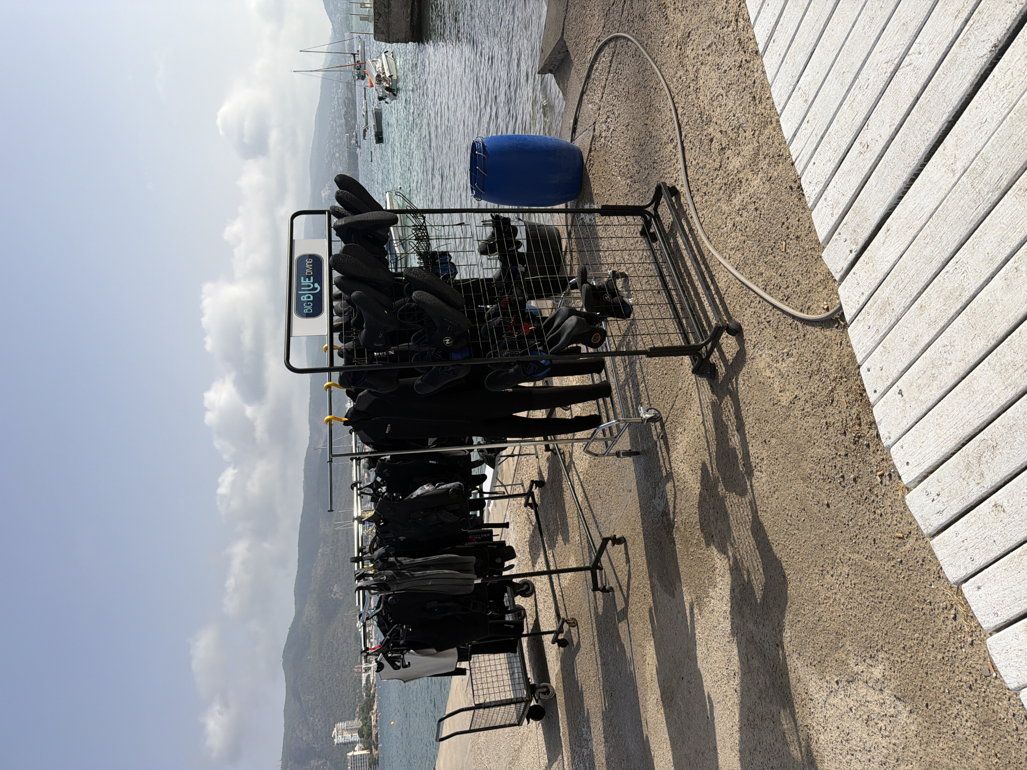 The image shows an outdoor scene in Calvià, Spain, likely a dive shop or a rental facility located near the water. In the foreground, a section of a white wooden boardwalk is visible. The main subject of the image is a collection of black wetsuits and other diving gear hanging on metal racks. These racks are mobile, with wheels. One rack has a sign with blue lettering that reads "BIG BLUE DIVING." To the left of the racks, there is a small wire shopping cart. A large blue plastic barrel is situated to the right of the racks. The background reveals a body of water with several boats and yachts. In the distance, there are hills with buildings and vegetation, characteristic of the coastal landscape of Calvià. The sky is partly cloudy. The lighting suggests it is daytime.
